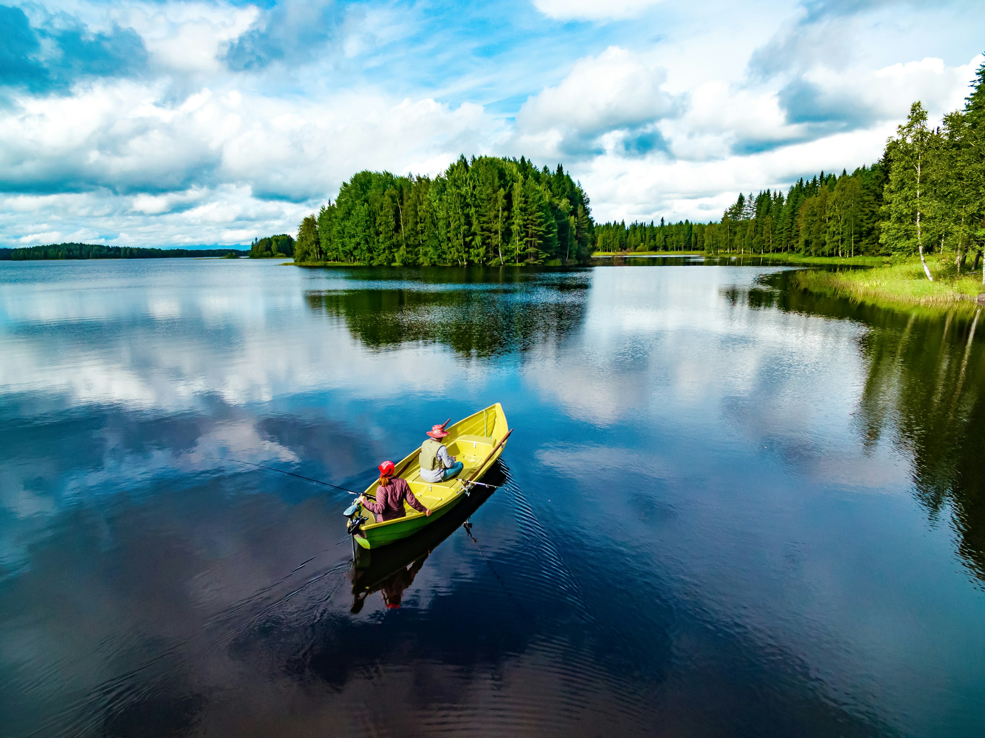 Aerial view of a yellow-pained fishing boat with young woman and man in blue lake in summer. Pine trees are seen along the coast, while the sky is partly clouded and reflected on the water