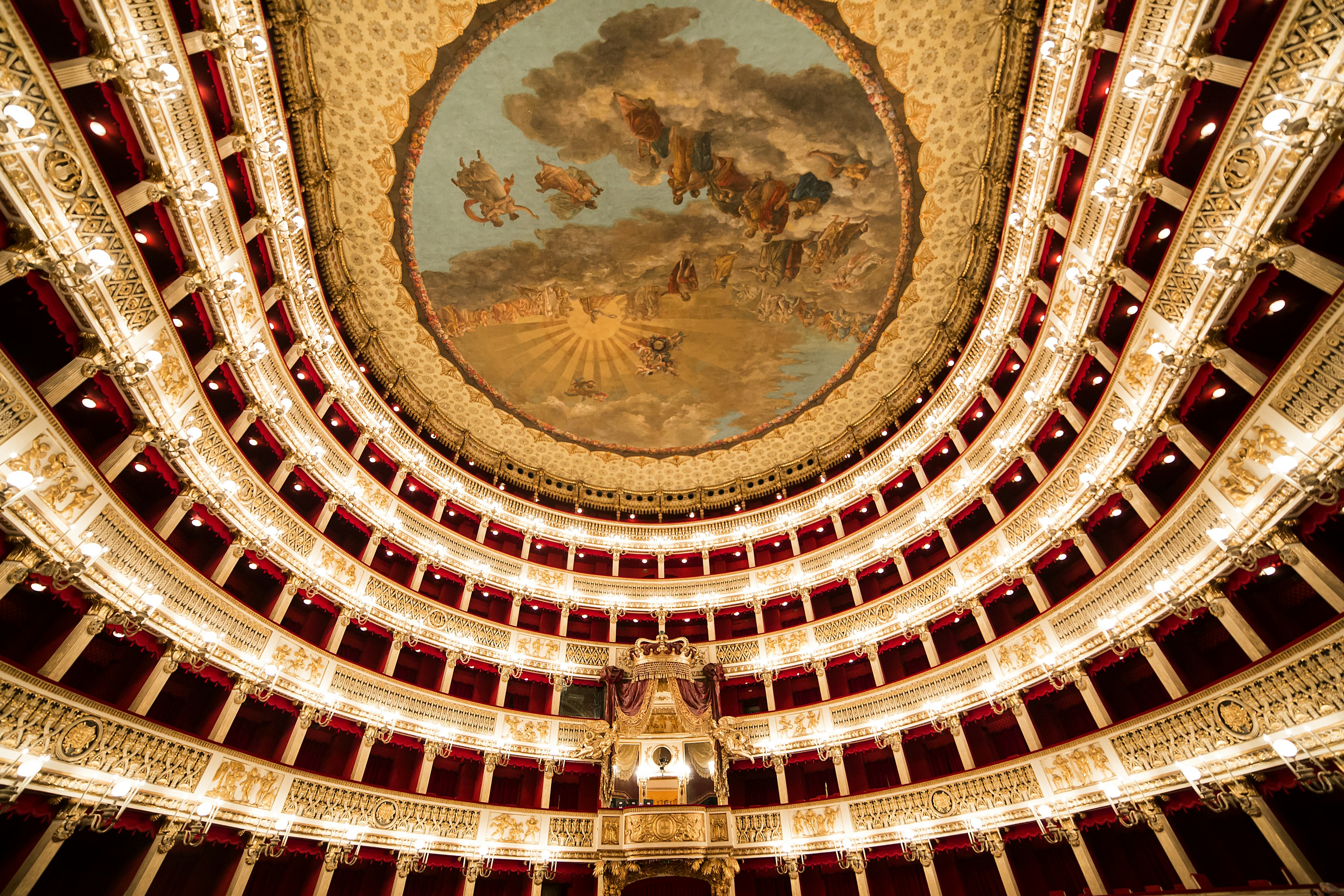 The interior of an opulent theater, with rows of private boxes and balconies, as well as the royal box with ornate decor. A painted fresco adorns the ceiling.