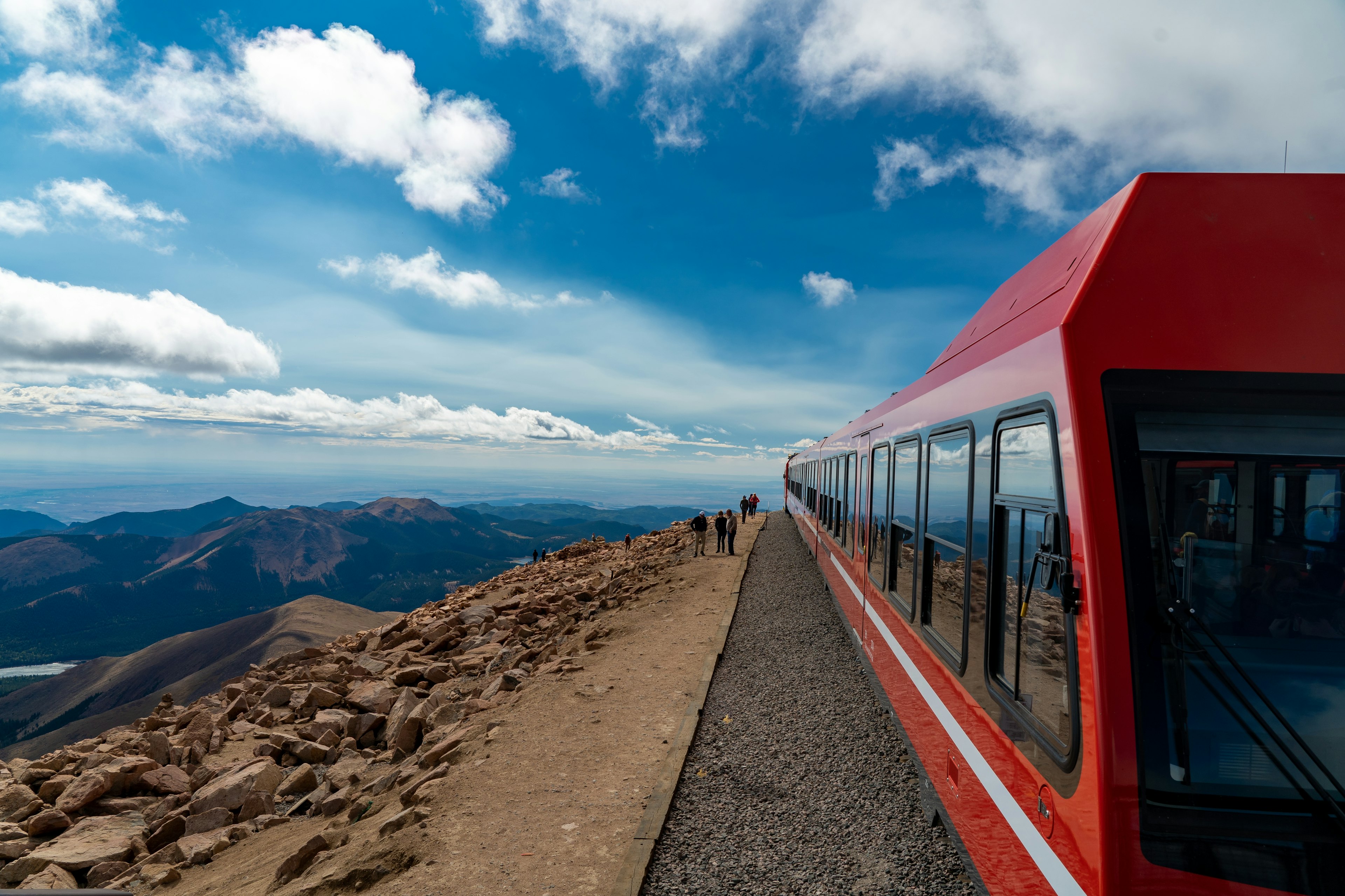 Red-Painted Train Cars Are Stationed at the Top of Mountain, with Passens Waiting to Board Along The Edge Of The Track. Dramatic Mountain Views are visible below the train.