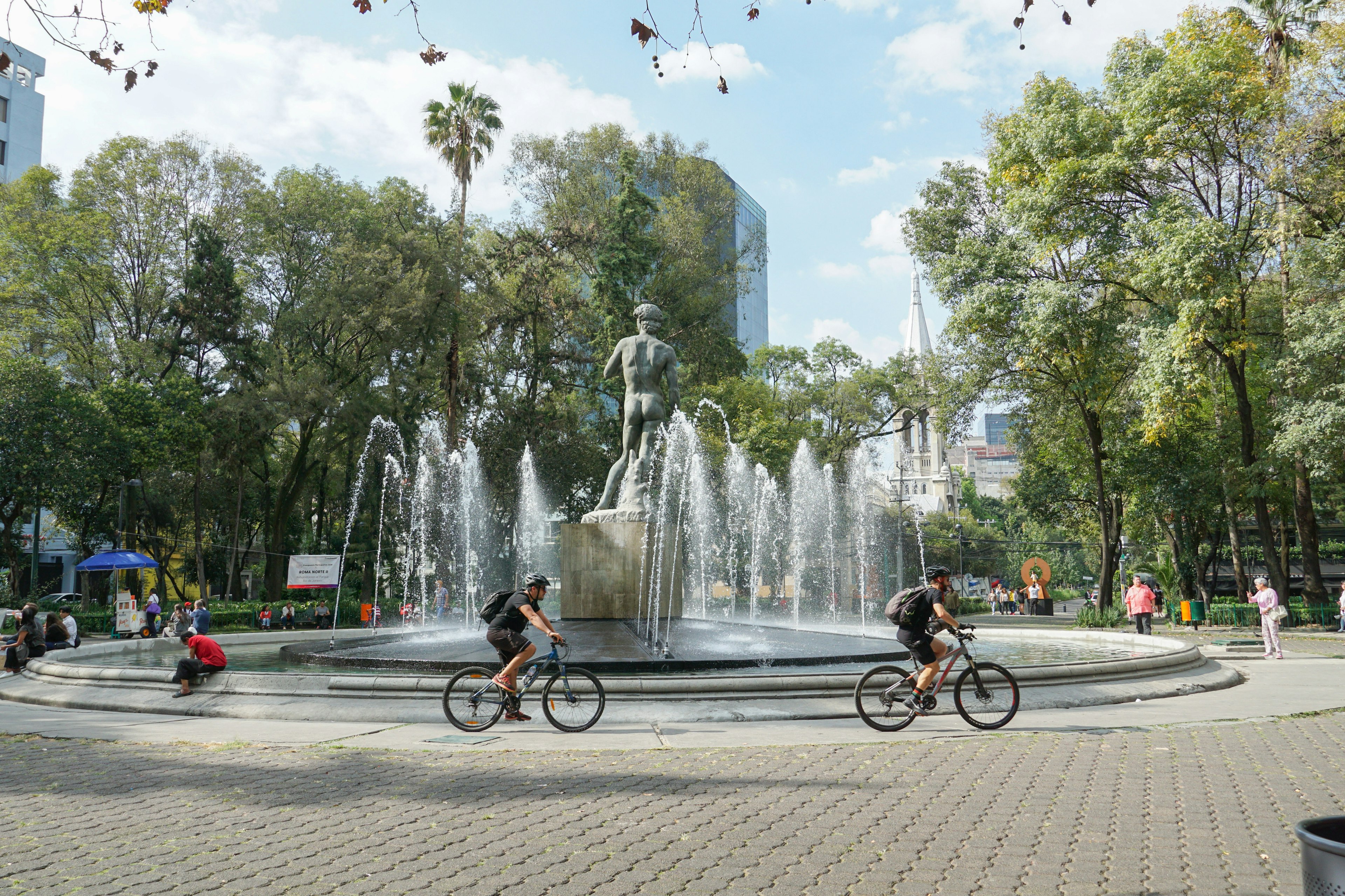 Young people riding their bikes around a large circular fountain with a replica of Michelangelo's David in the center in Plaza Rio de Janeiro, CDMX.