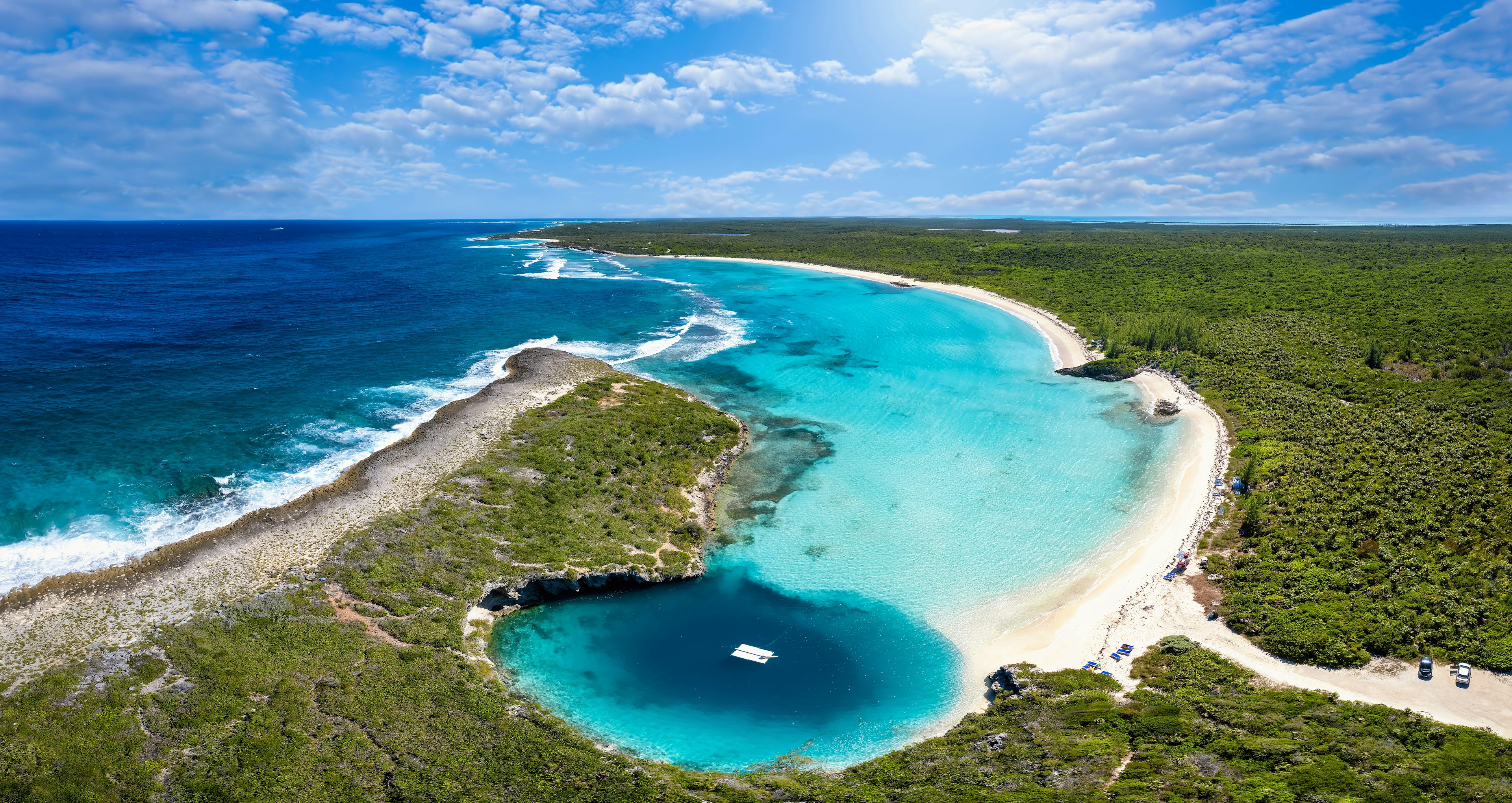 An aerial view of a sliver of land with greenery that separates the coastline from a lagoon that contains a dock in the center of a blue hole in the Bahamas. The water has areas of contrasting deep blue and turquoise, and a white sandy beach curves out from one end of the cove.