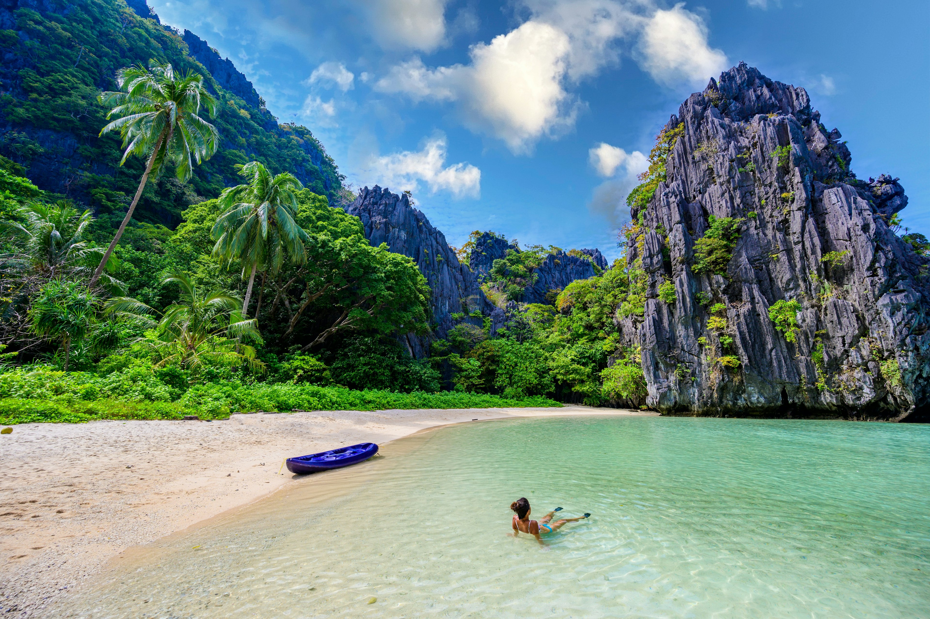 A woman floats in the shallow waters at a tropical beach. She is alone; karst towers and palm trees are available on the shore