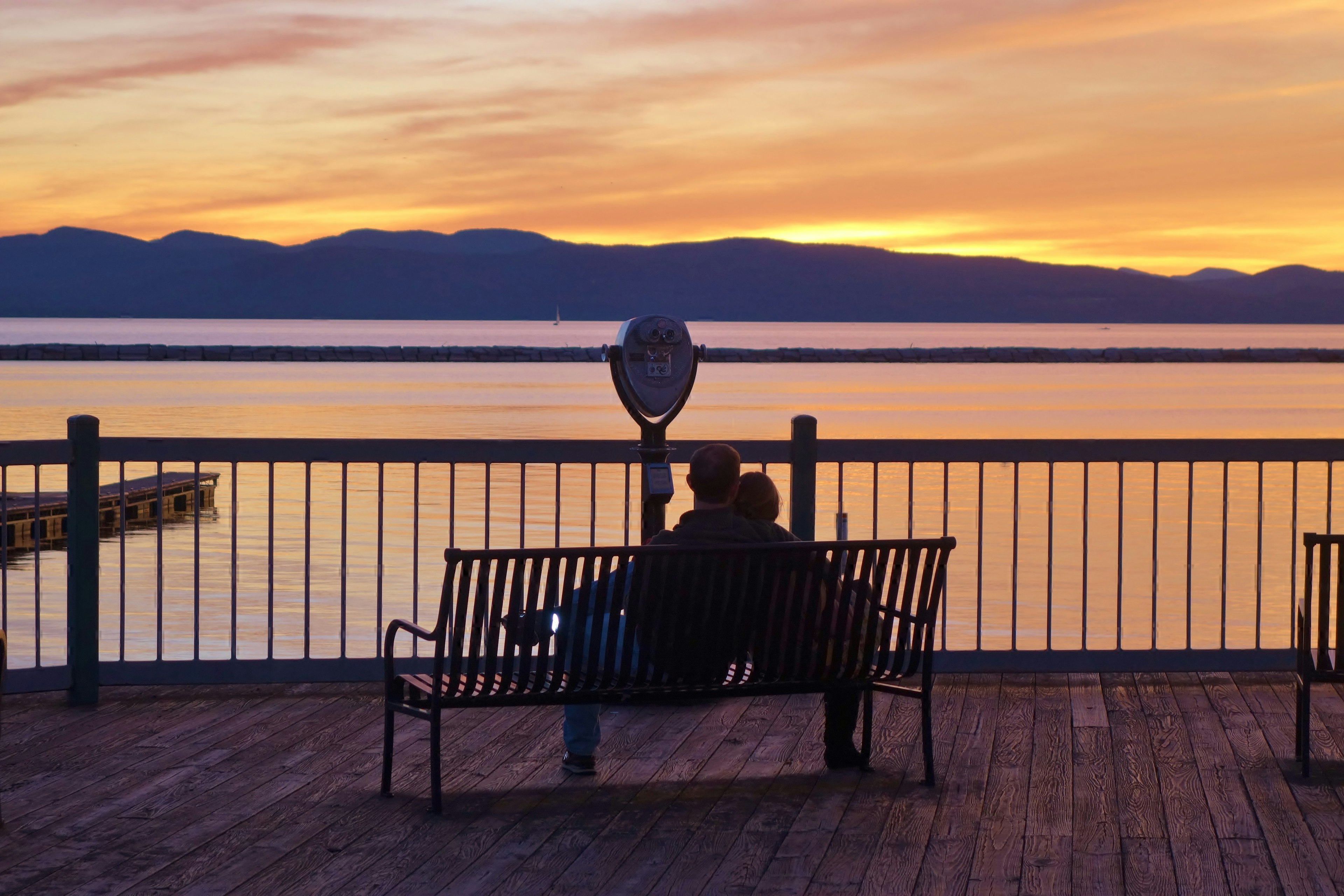 Watching an amazing Lake Champlain sunset a couple sitting on a park bench is hugging as the sky glows n orange over the Adirondack Mountains at the end of a beautiful fall day in Burlington, Vermont.