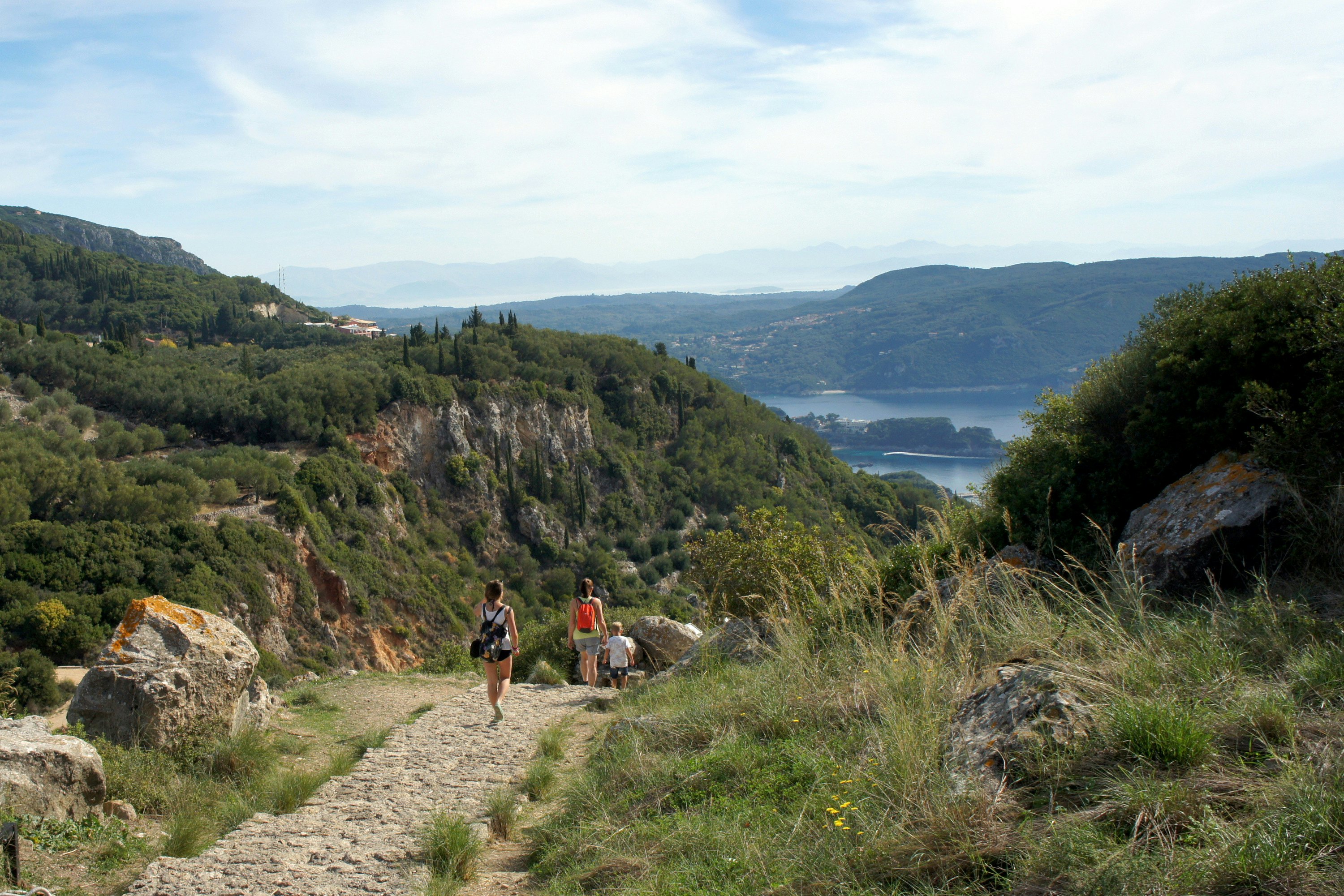 Hikers carry small packs along a cobbled path through scrubland down towards the coastline.