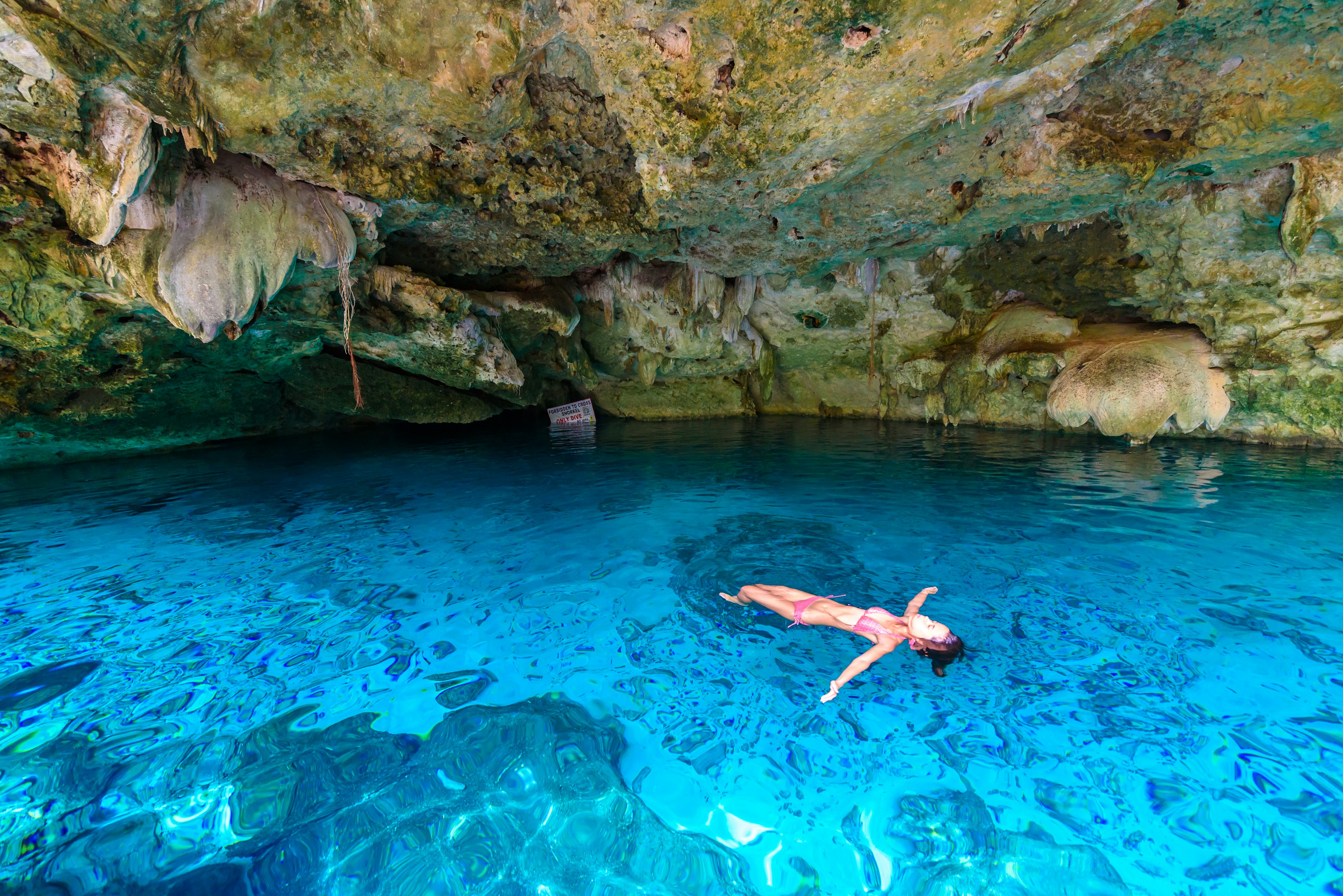 A woman floats on her back in the blue waters of a cenote. The rock formations of a cave are visible above her.