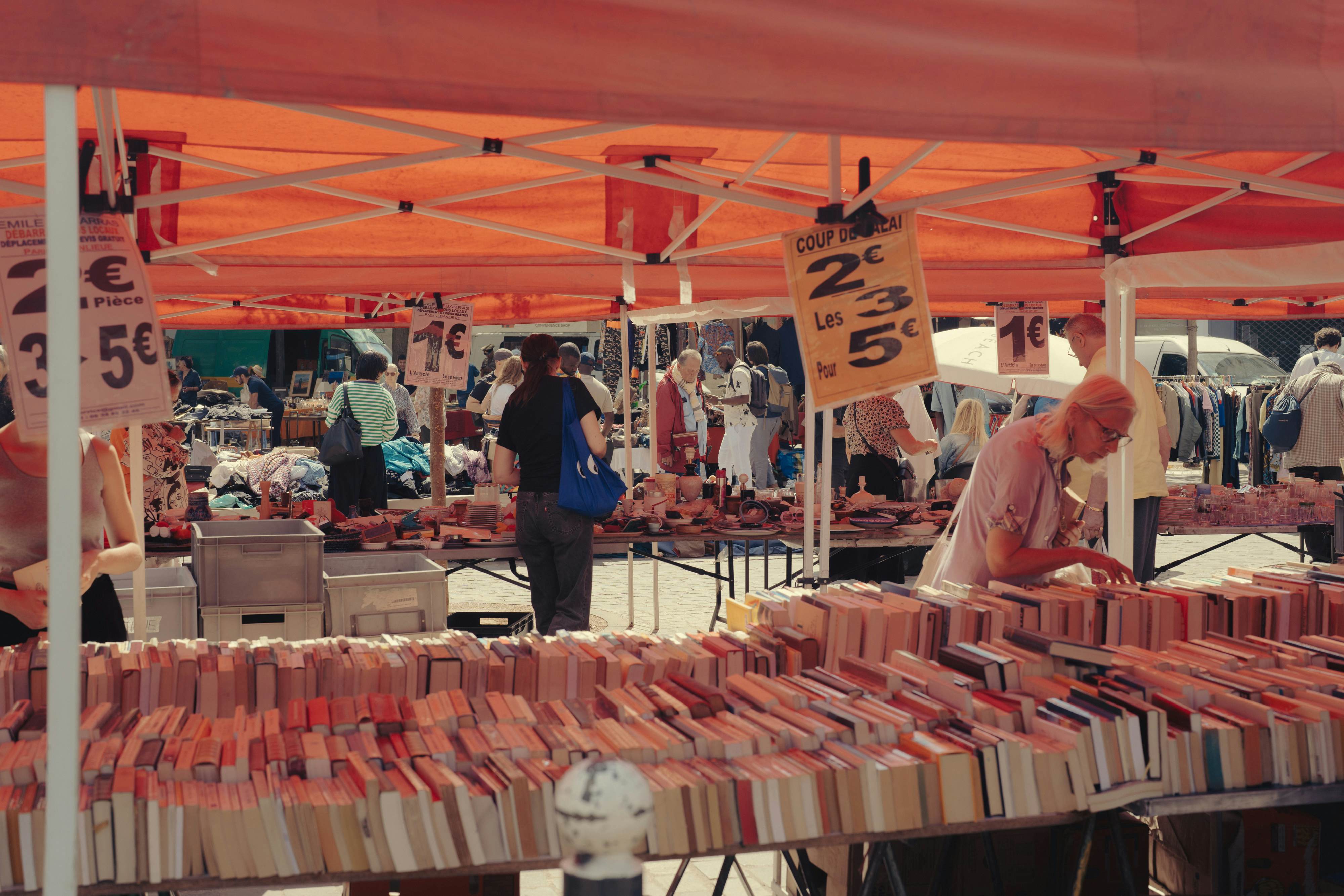 People browse books at a stall with a cover at an outdoor market in a city.