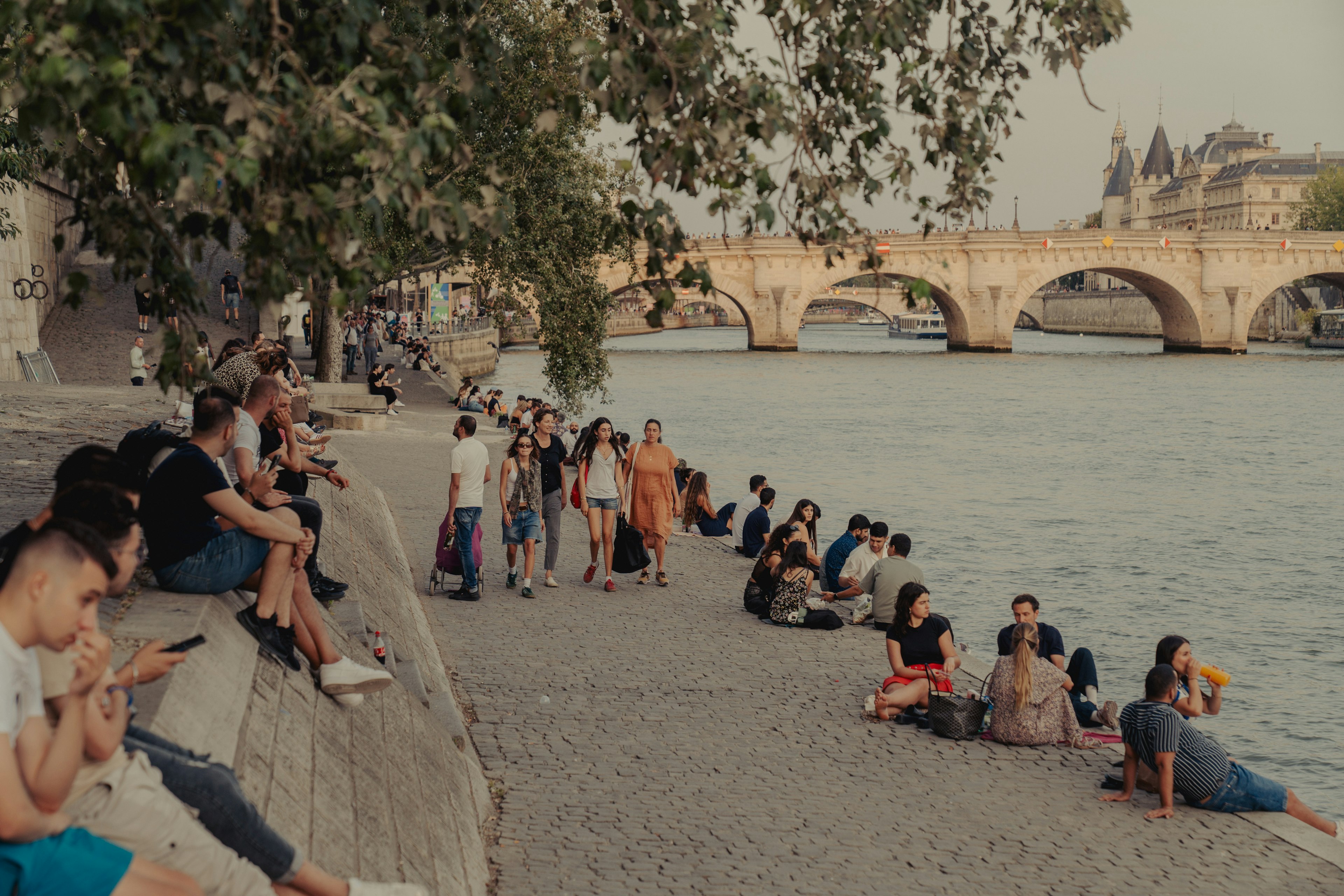 People relax by a river with a large stone bridge crossing it