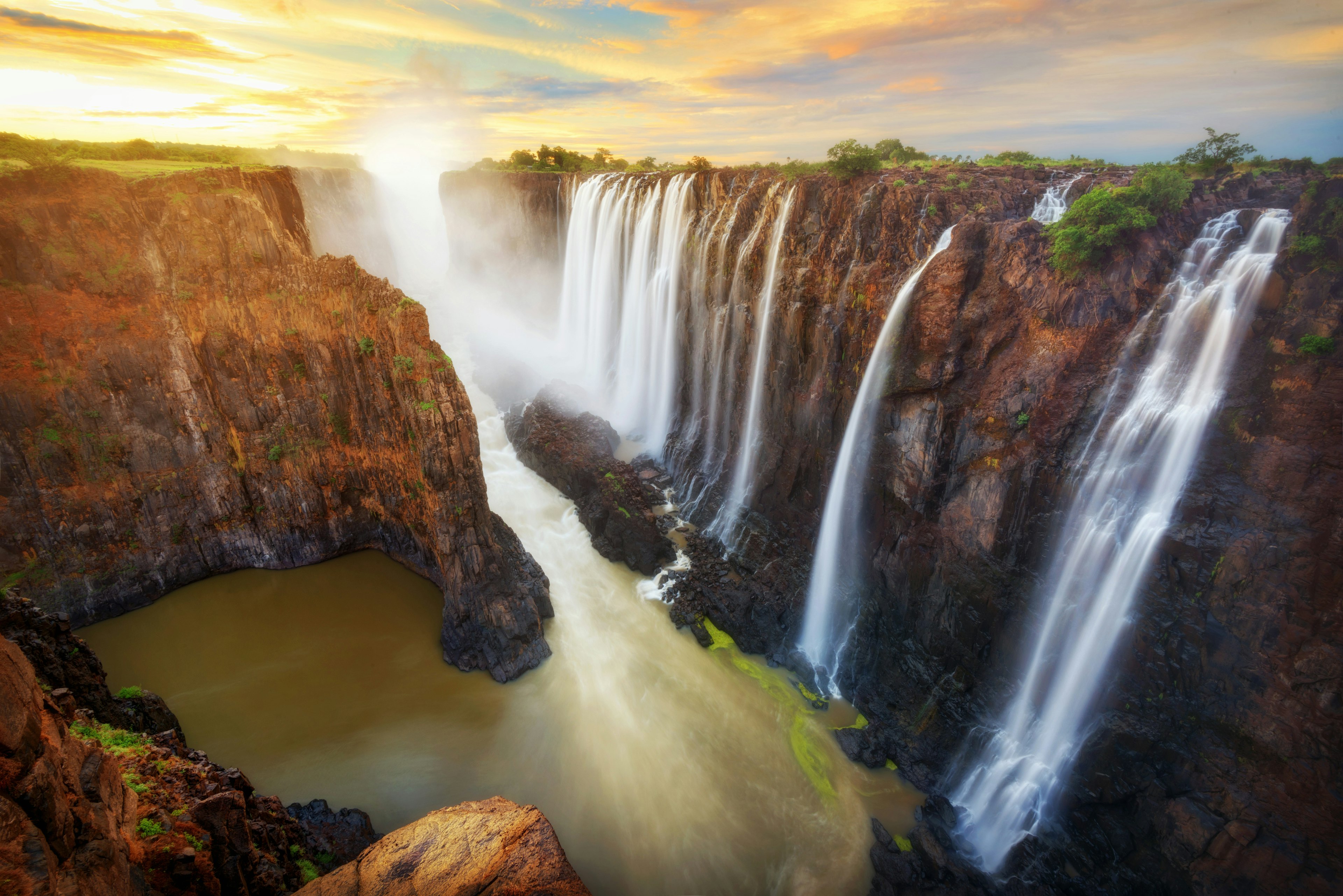 A vast series of waterfalls plunge over rocks that glow red in the sunset