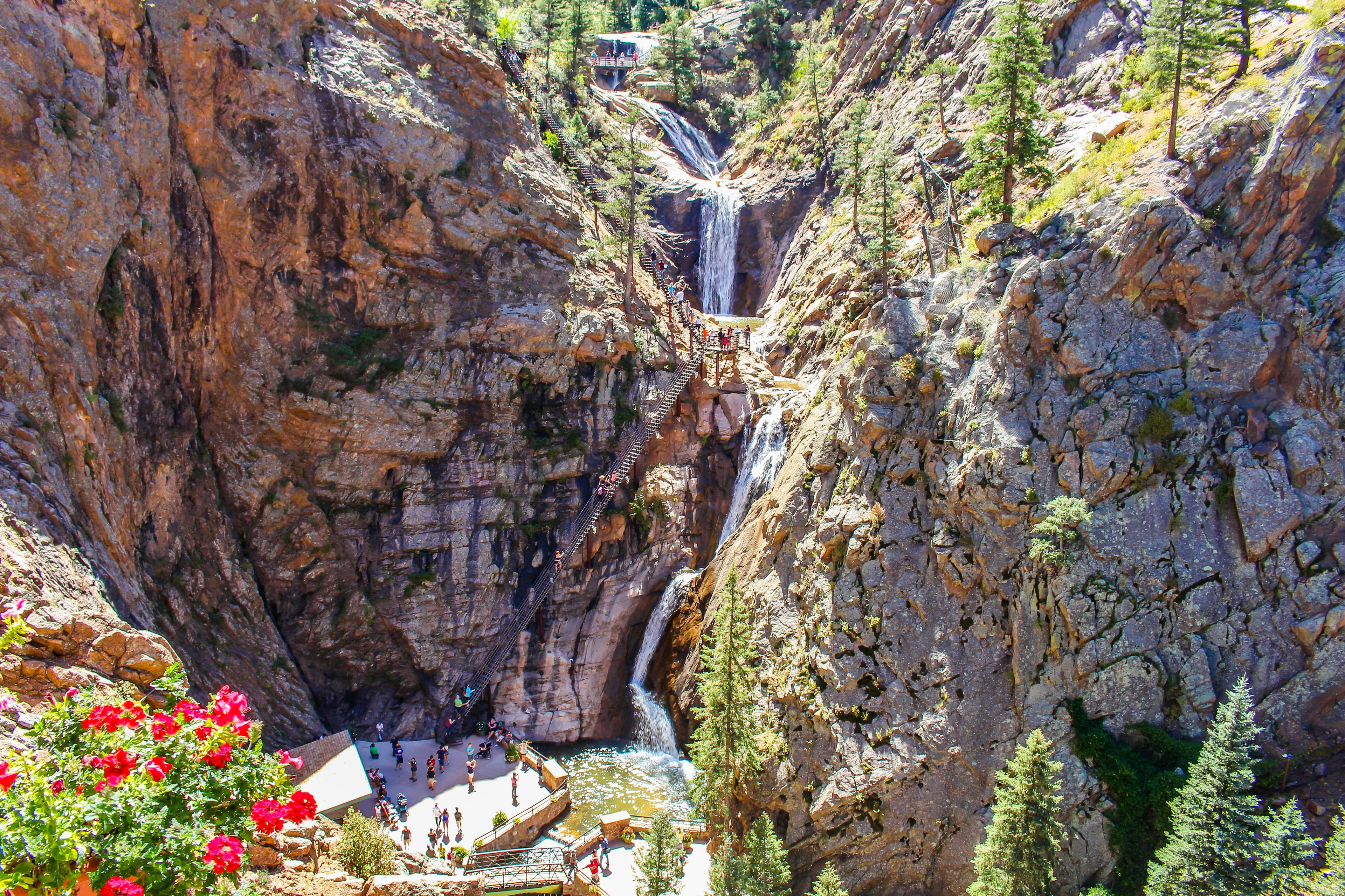 A wide view of a set of waterfalls that cut through a rocky face. Groups of visitors are visible at an observation deck below, and a bridge over the midpoint of the falls.