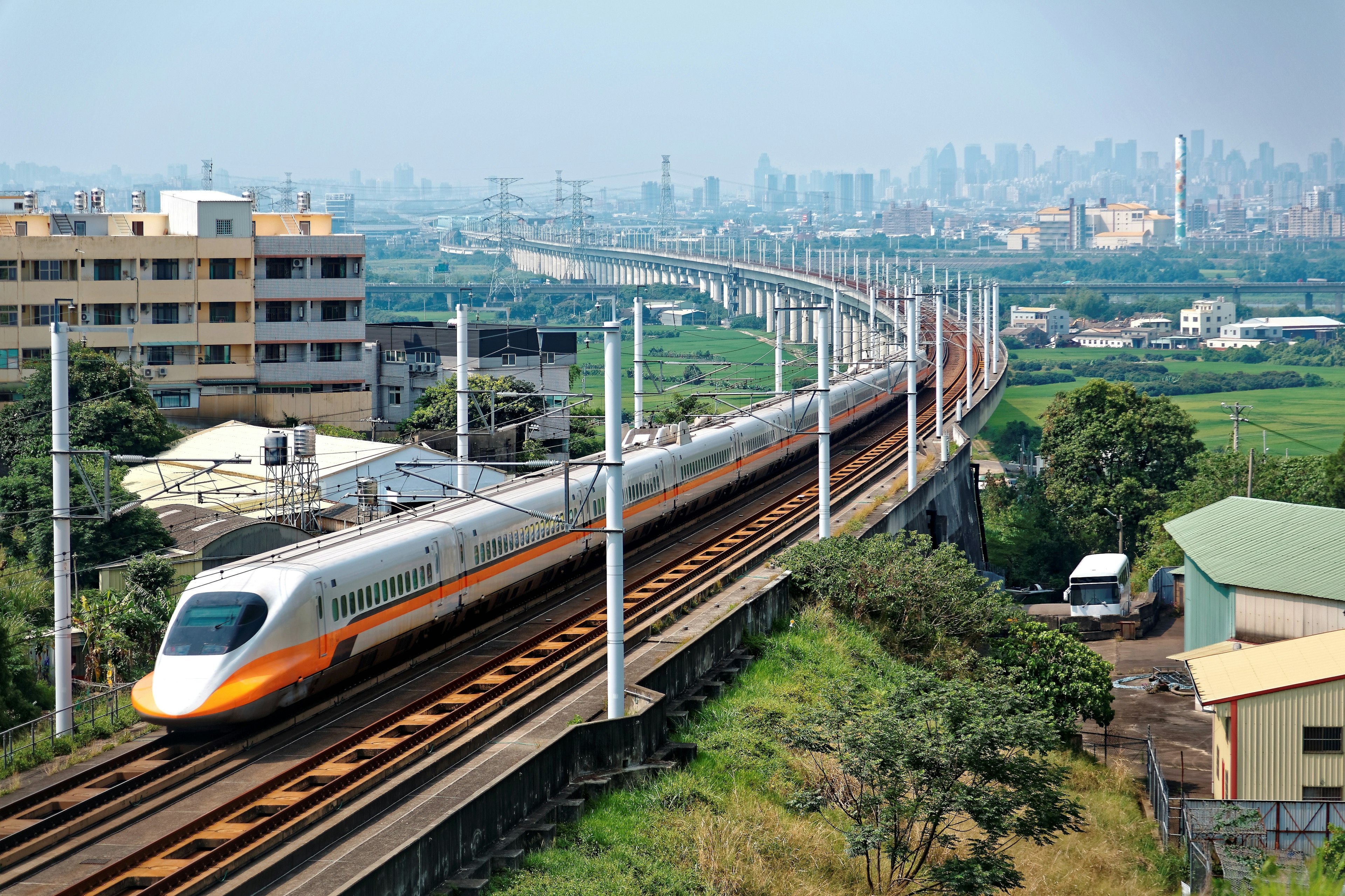 A white train with an orange stripe departs from a large high-rise city