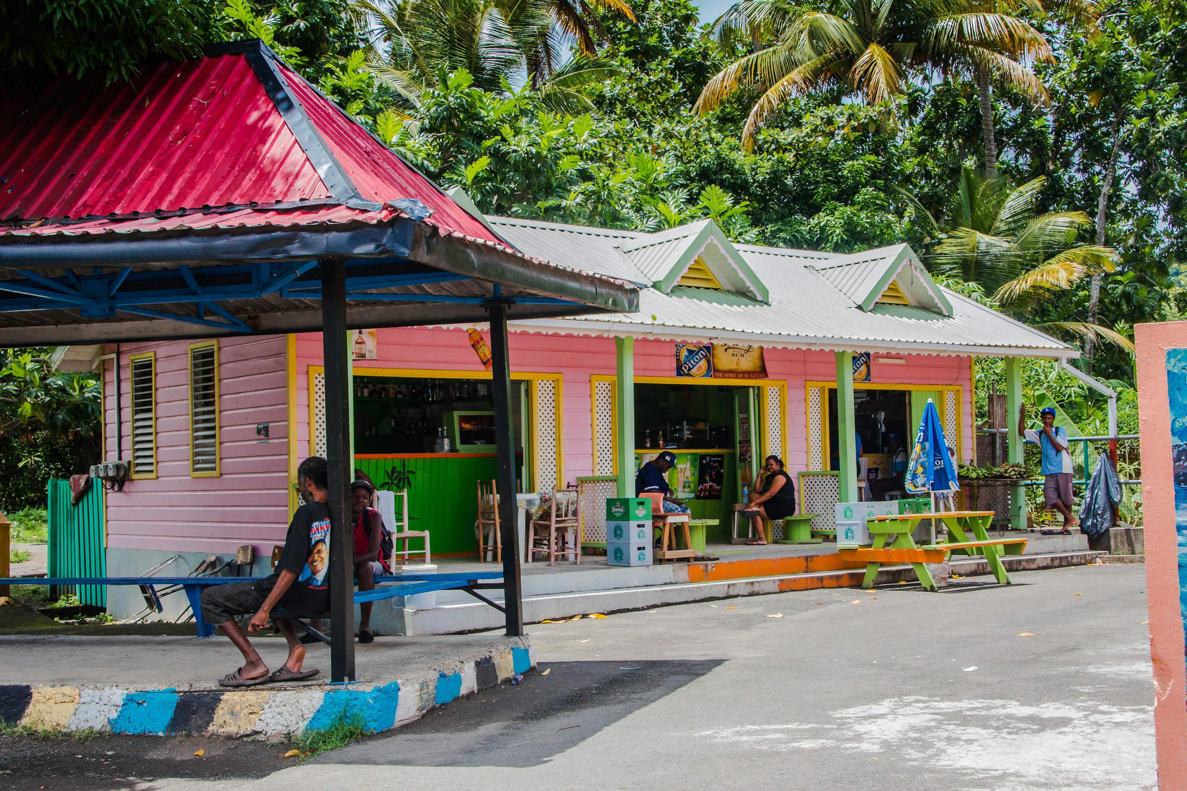 People sit on benches on a porch and pavilion in front of a bar in a tropical locale. The building is painted pink, with green accents.