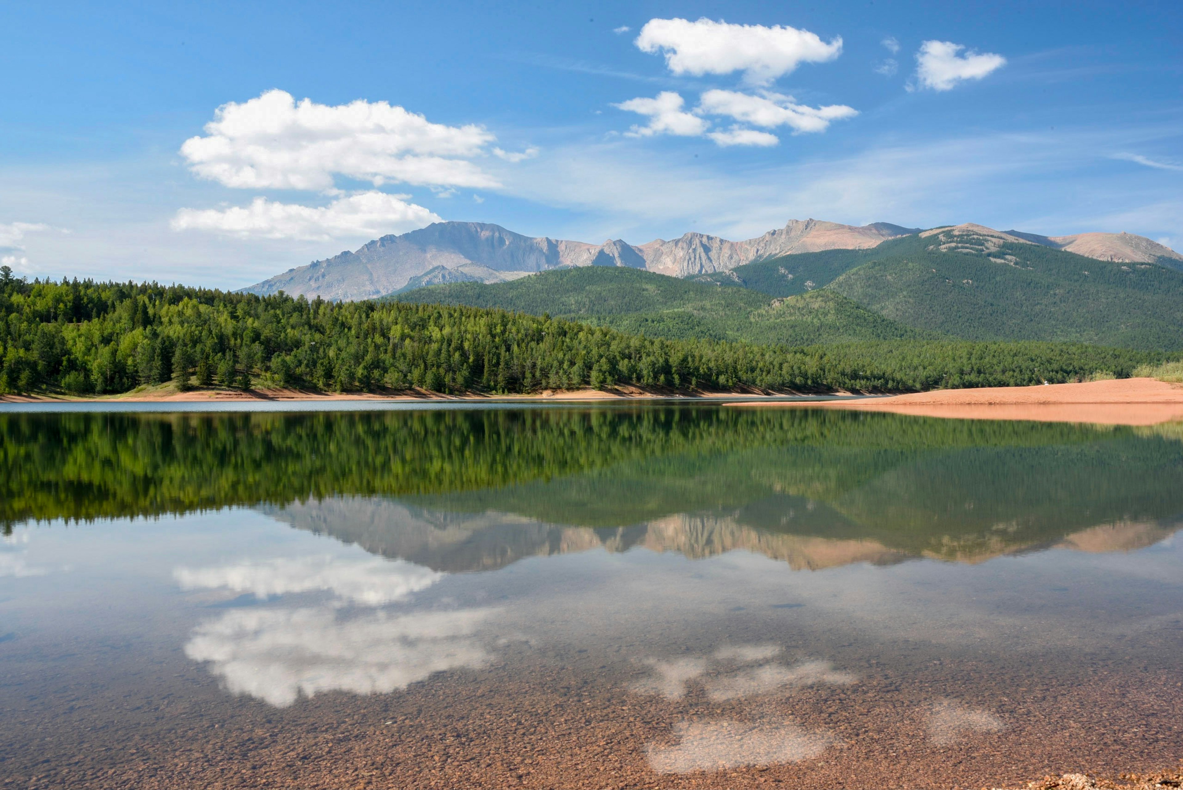 A Blue Sky and Clouds Are Reflected in a Still Mountain Lake. Pine Trees Are Visible Along The Shore, and Mountains Can Be Sea in The Distance.