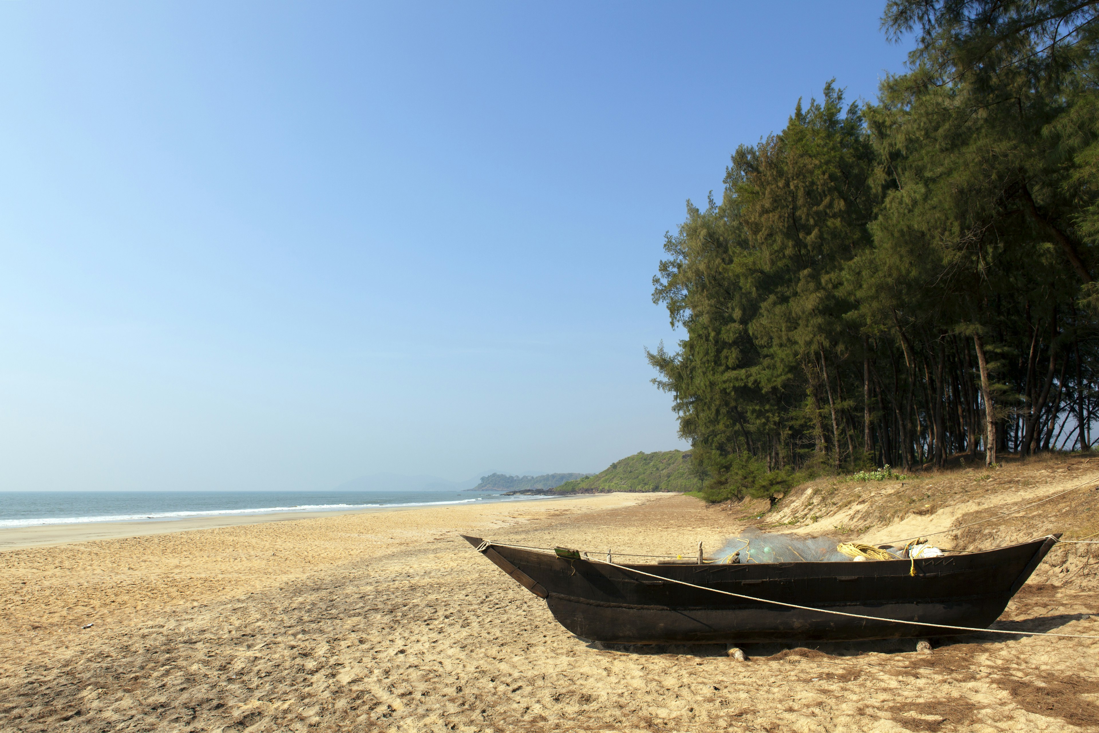 A small wooden boat sits on an empty beach near a line of trees.