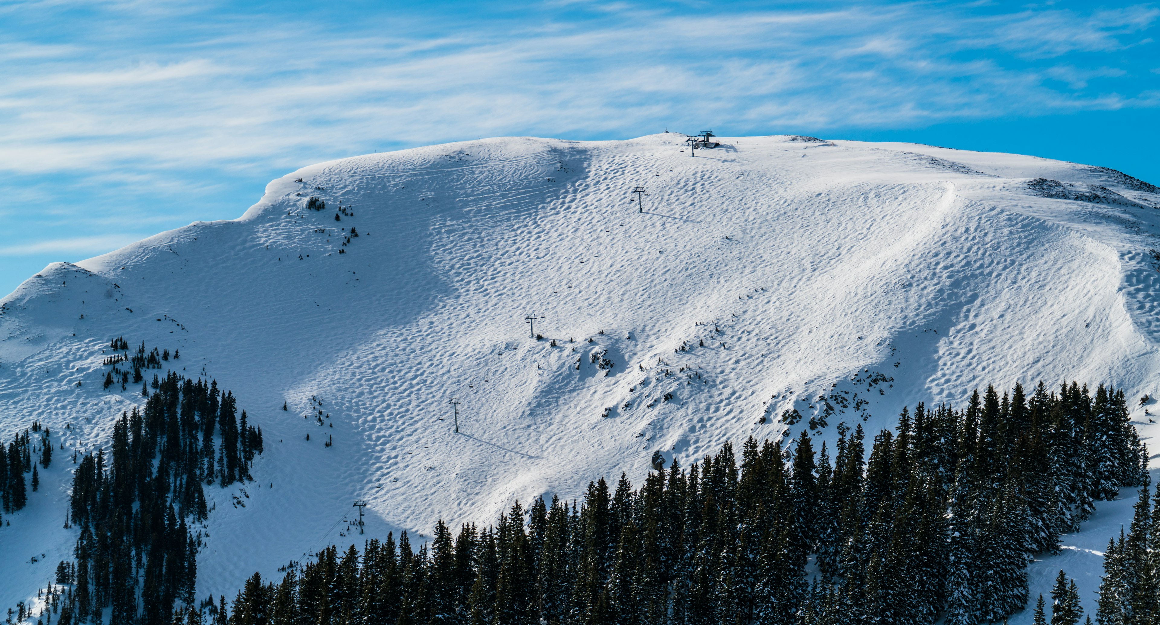 An aerial view of the peak of a snow-covered mountain, with pine trees and the towers of a chair lift for skiers leading to the top.