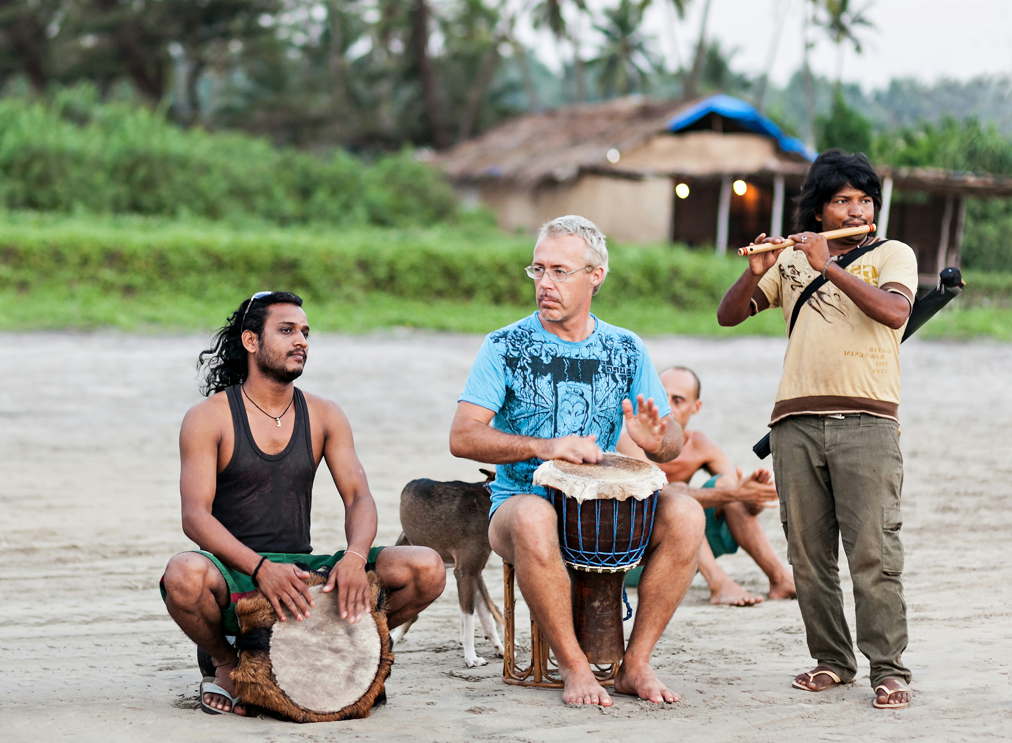 A group of men playing drums on a beach. A structure and palm trees are visible in the background.