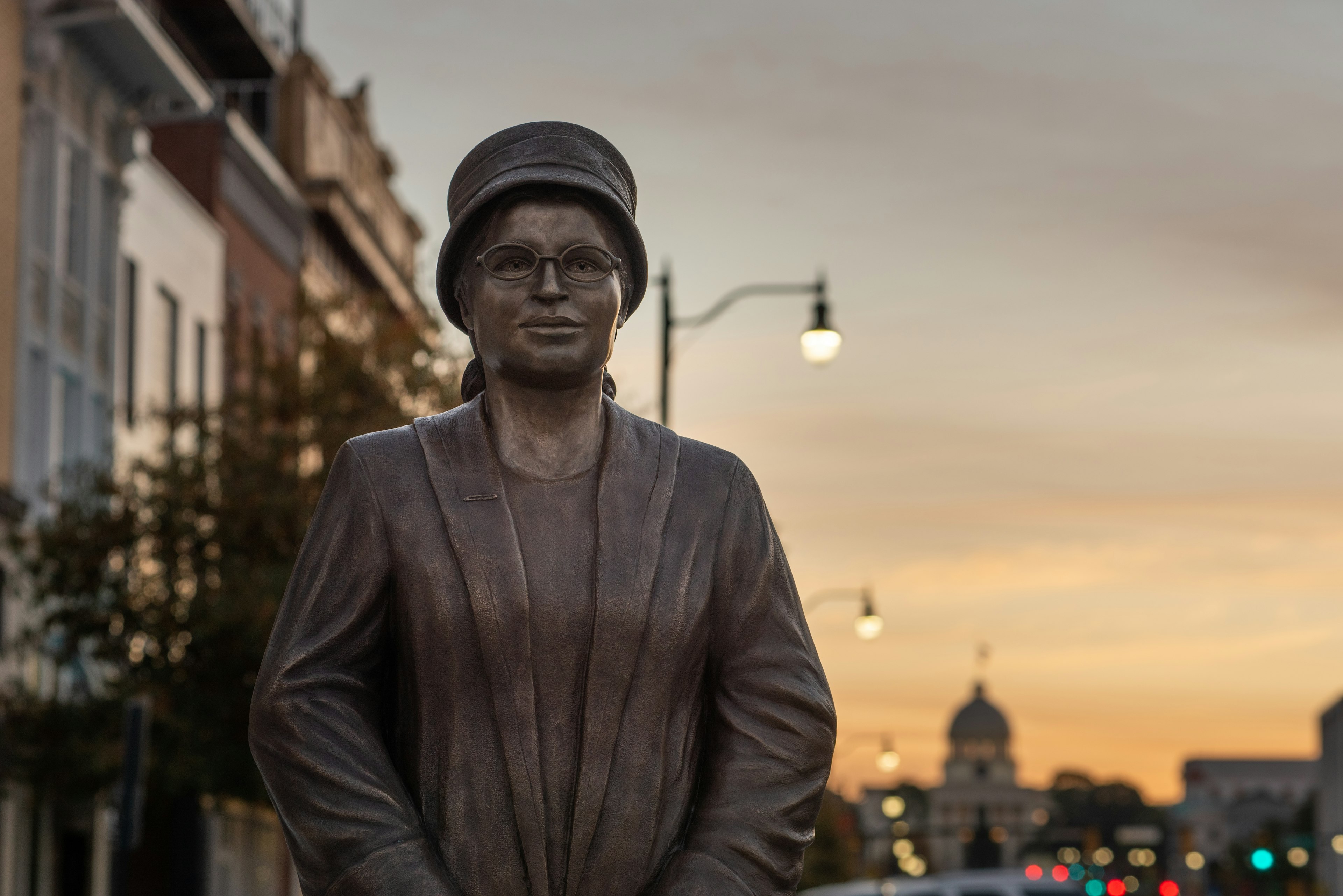 A life-size sculpture of Rosa Parks on a street in Montgomery, Alabama, USA. Dusk light is seen in the sky, with the state Capitol of Alabama silhouetted in the distance.