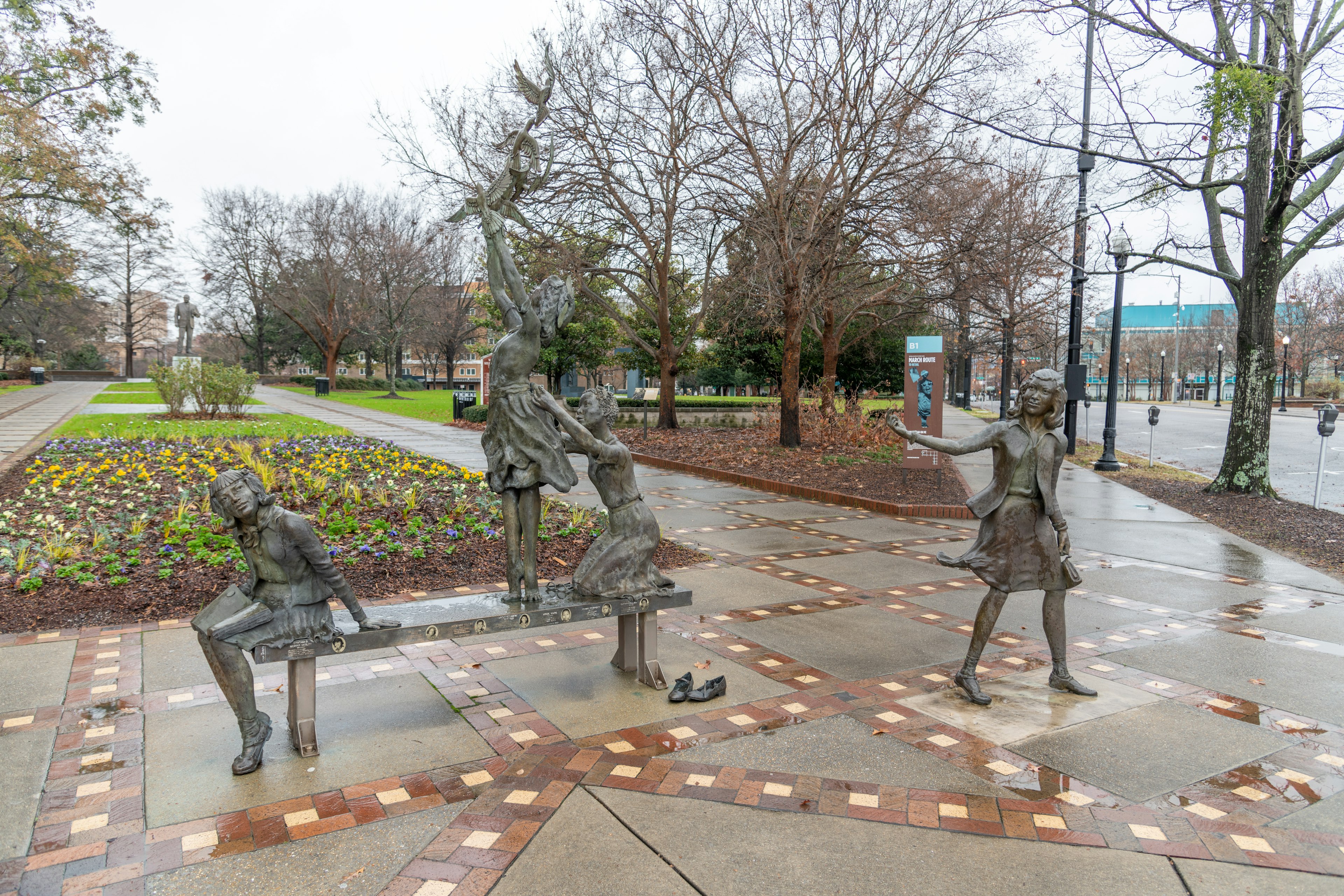 A sculpture in a city park depicts four girls playing. The sidewalk is wet with rain.