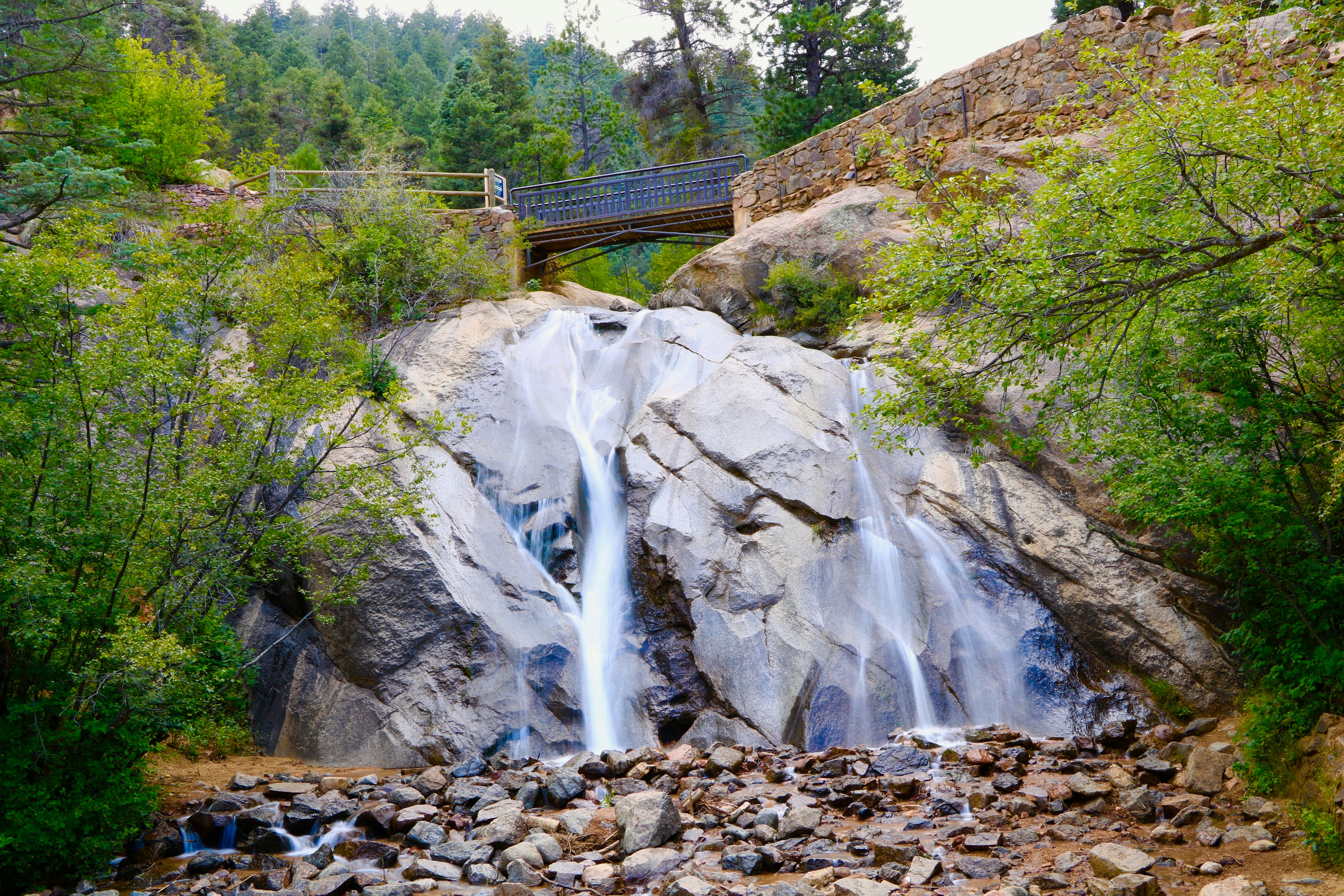 A view from below of a beautiful waterfall spanned by a bridge at the top, surrounded by green vegetation