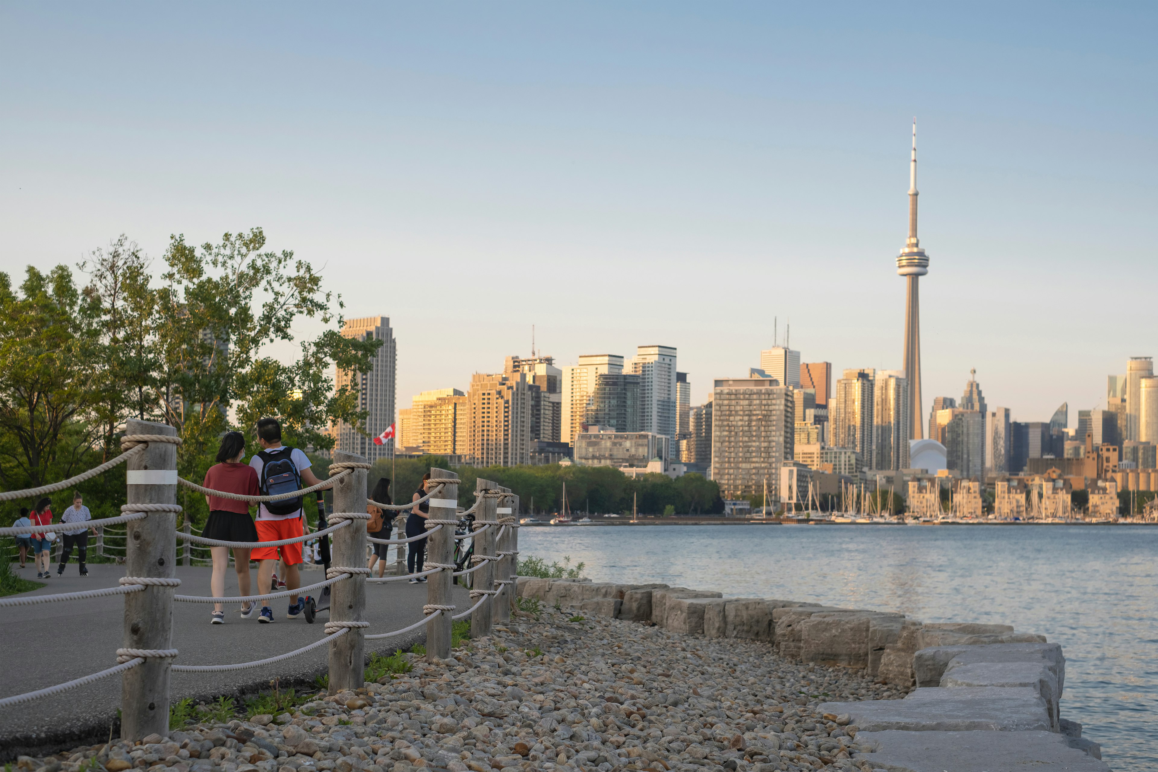 People walk and scoot along a lakeside path with a view of a city skyline