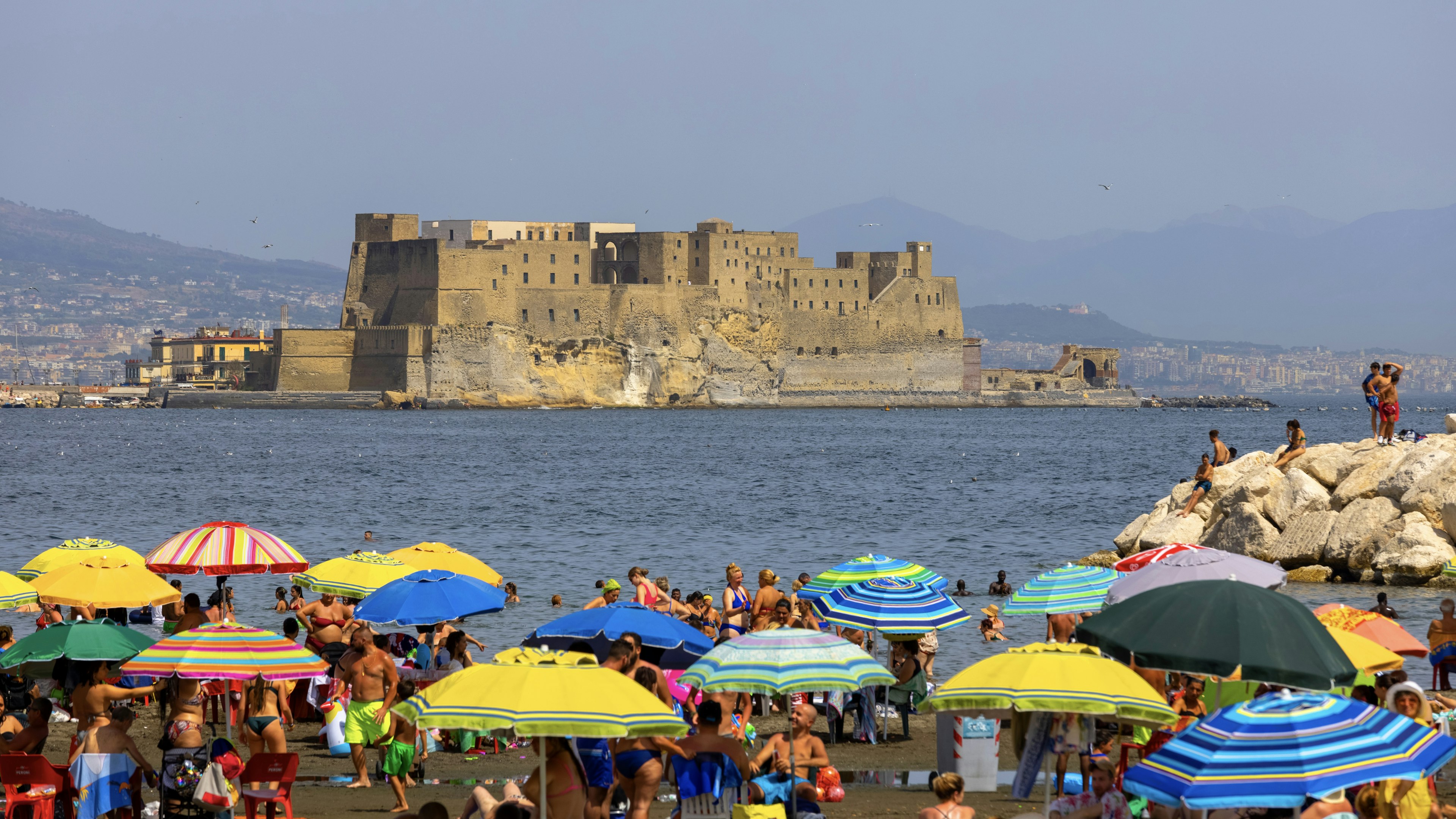 People relaxing on the Mappatella Beach by the Tyrrhenian Sea next to the boulevard Chiaia, colorful umbrellas, Castel dell'Ovo in the background.