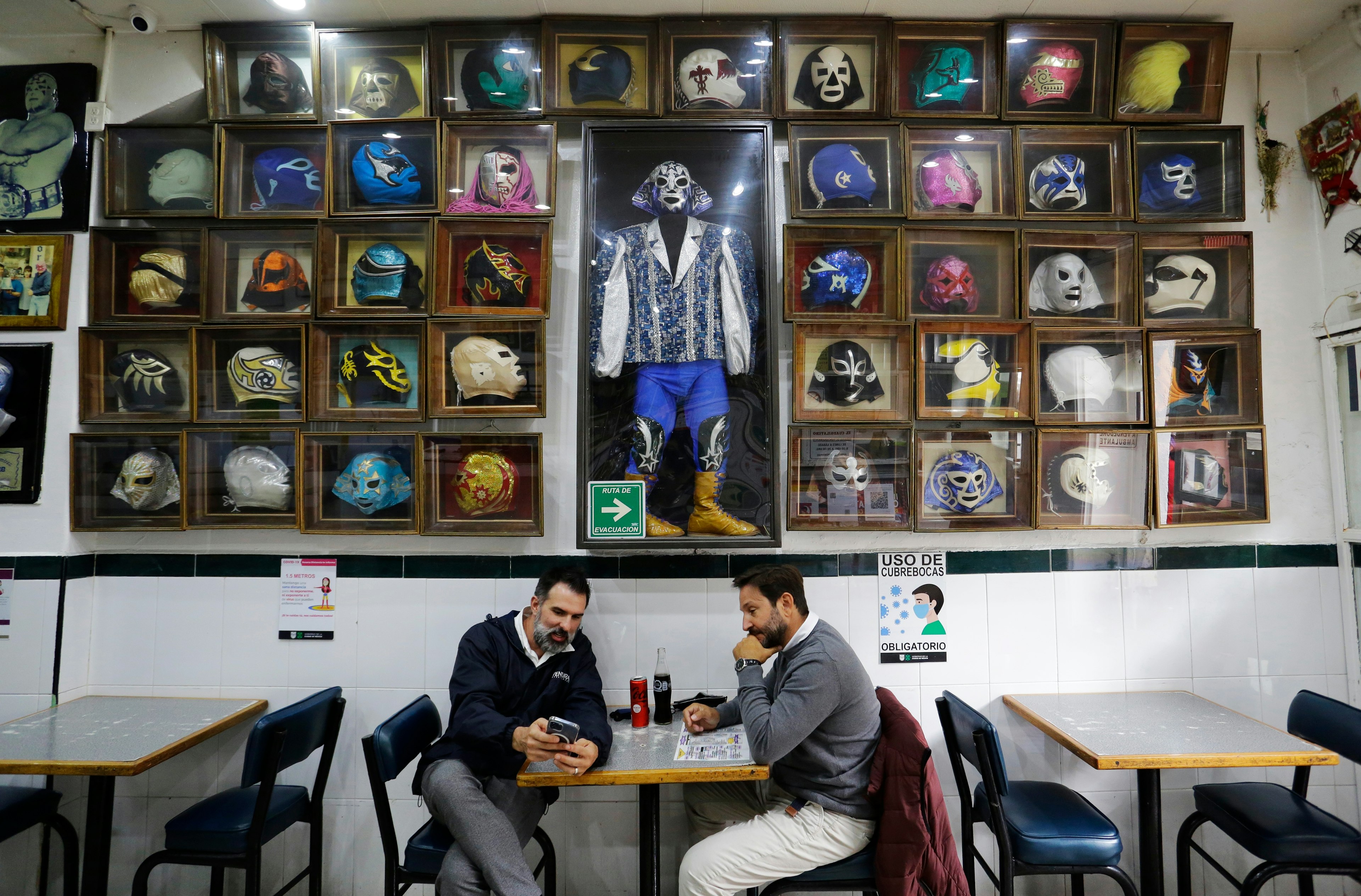 Two men look at a phone at at table in a sandwich shop under a wall covered with colorful masks of Mexican wrestlers