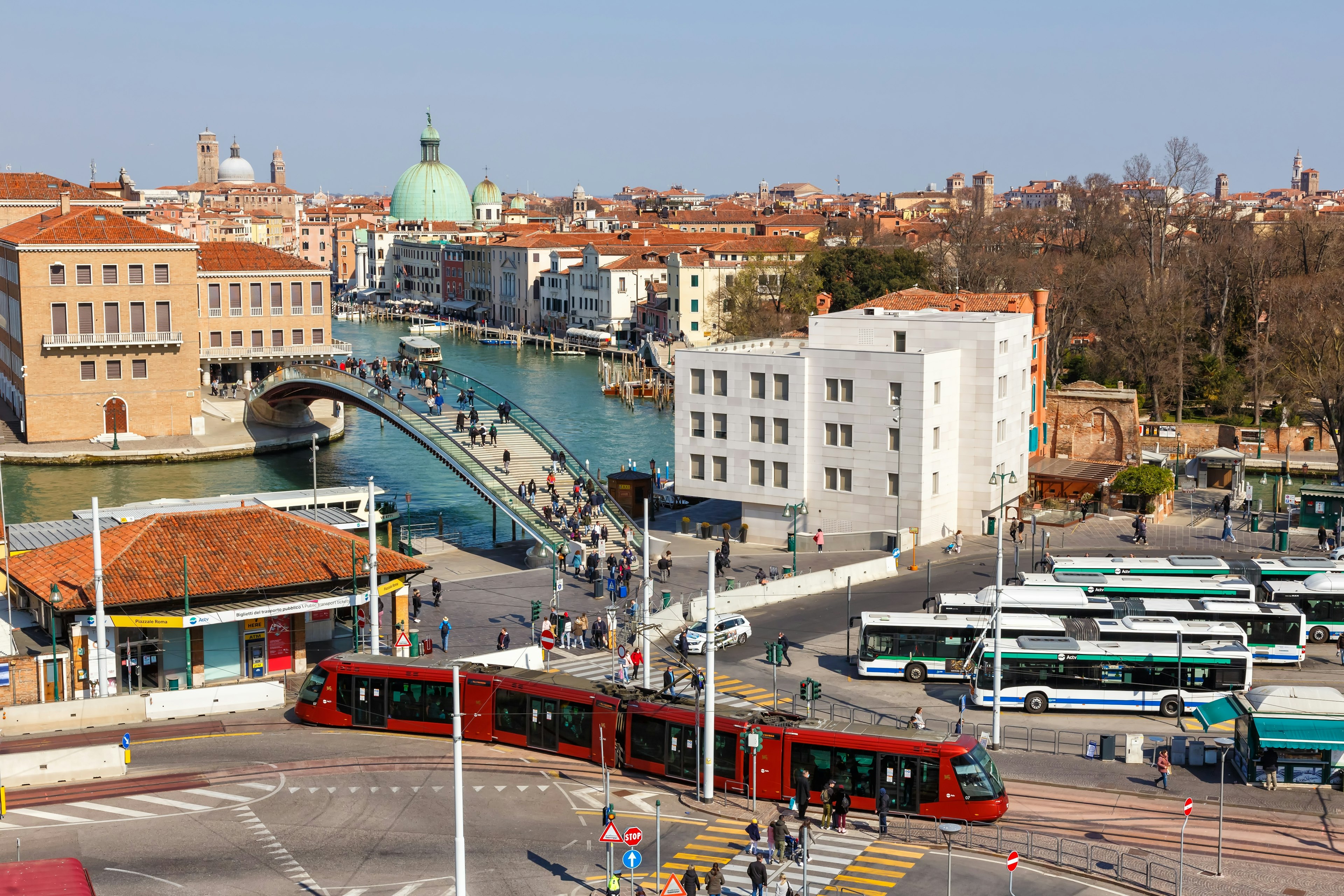 An aerial view of a plaza with a tram, buses and a bridge over a canal, with rooftops and church domes in the distance