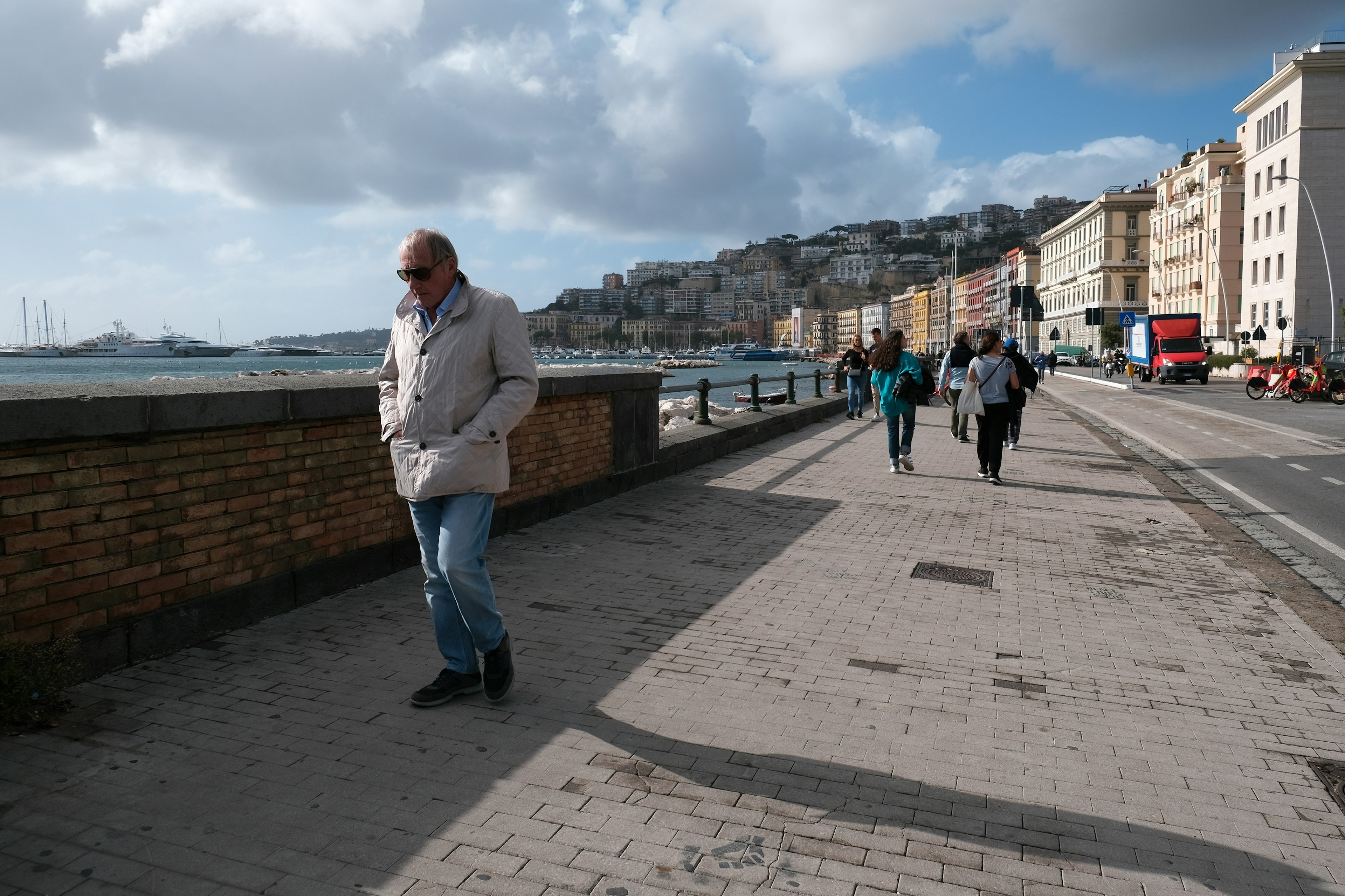 People walk in cool weather along a waterfront sidewalk in a city. The sun illuminates apartment buildings across the street in the distance.