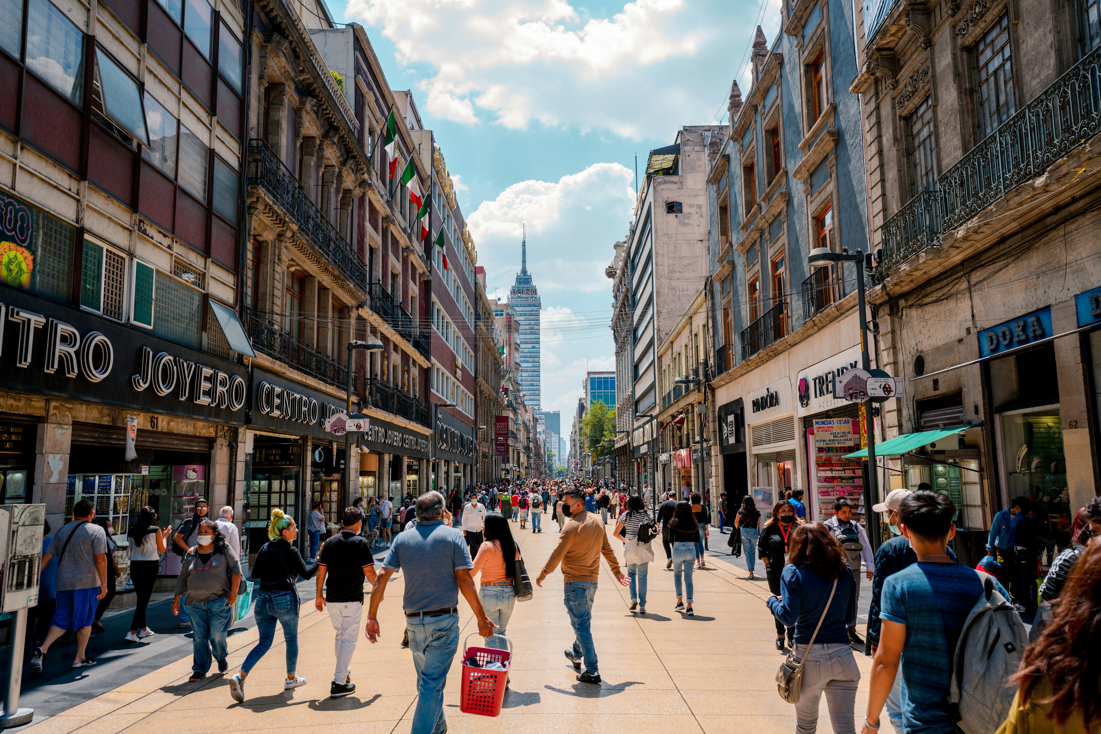 People walk on a busy pedestrianized street in a big city. Shops in buildings in a range of architectural styles are open on either side of the street.