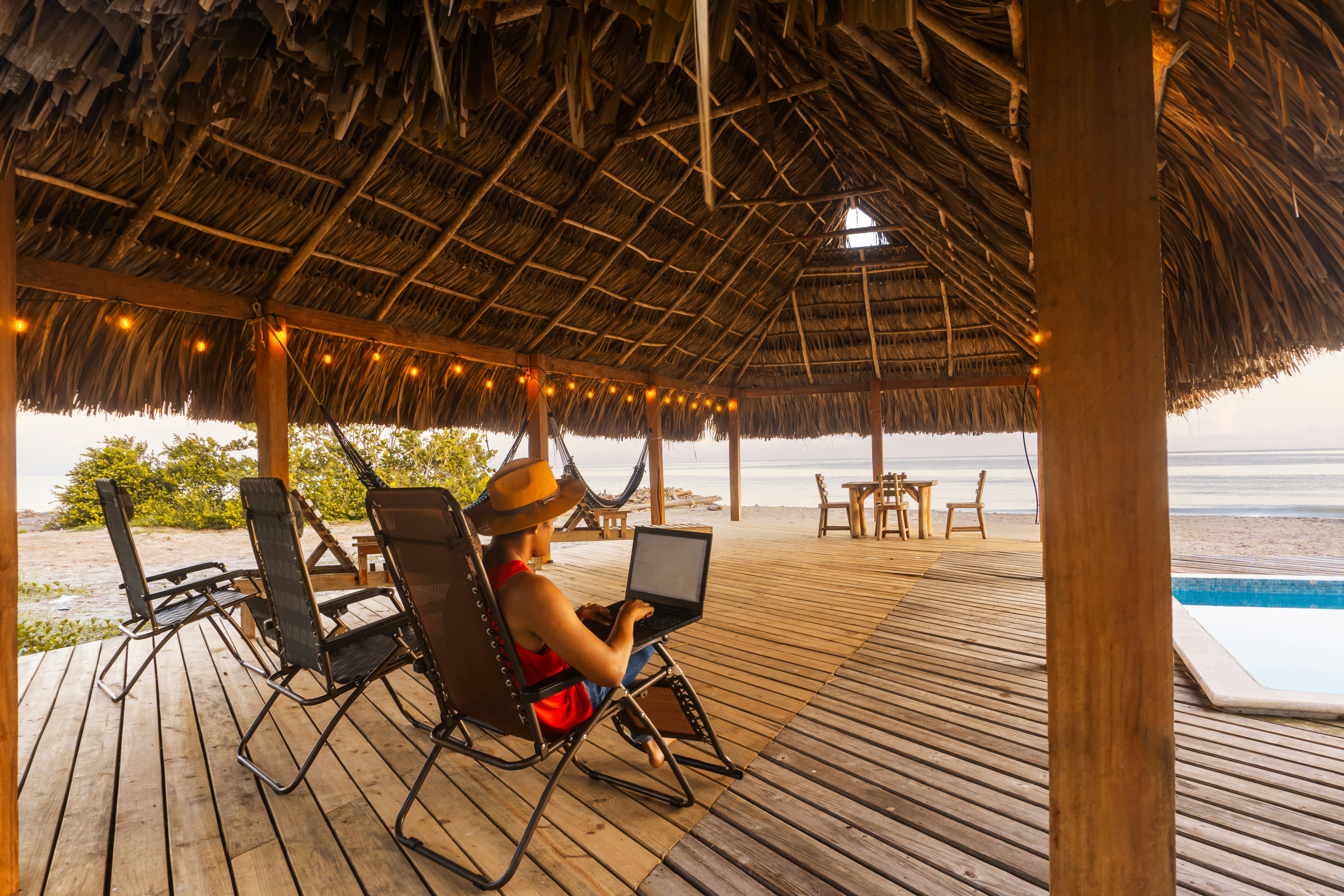 Man with a hat sitting on a chair in a thatched hut and working on a laptop. The beach is visible just beyond the covered platform.