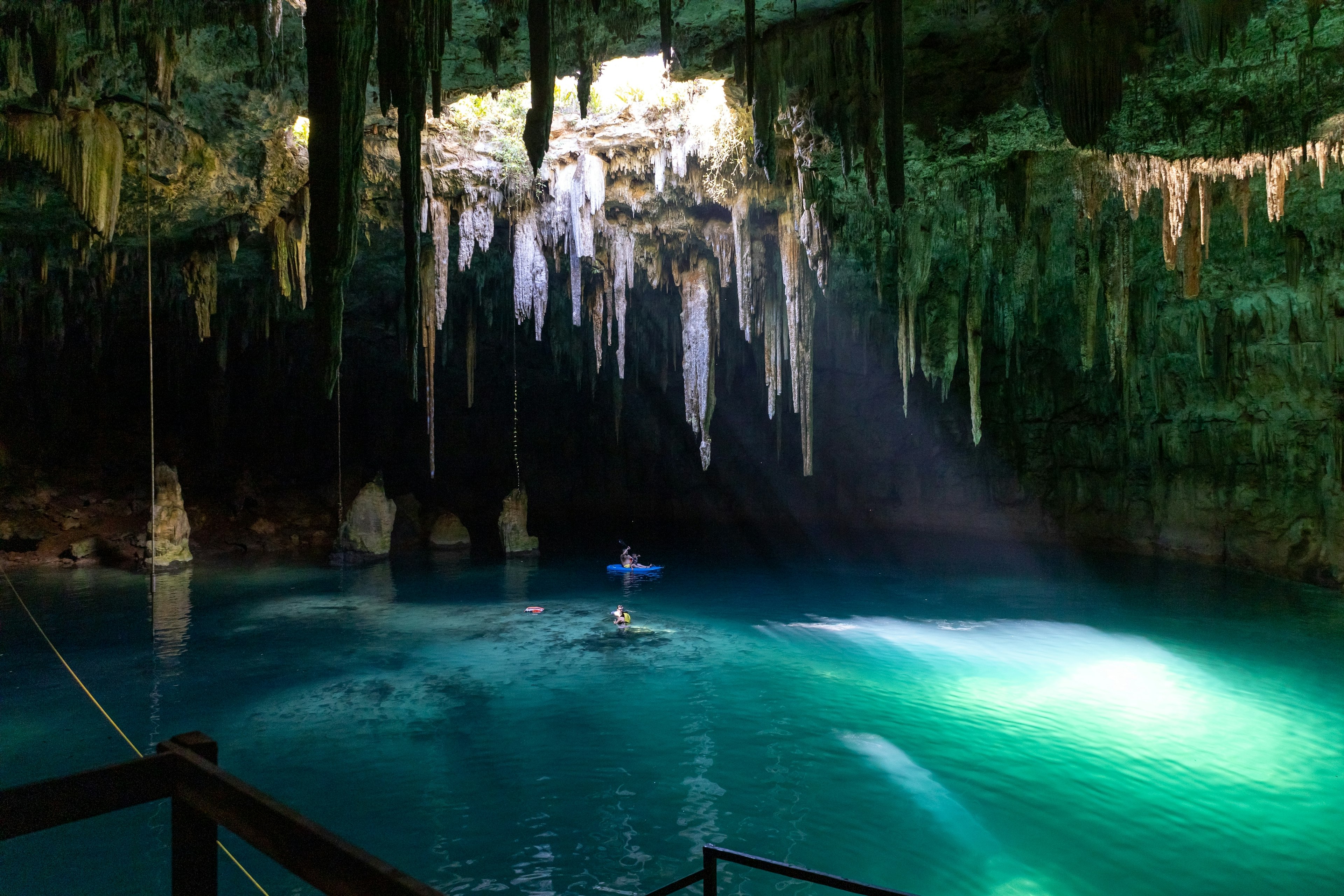 People swim in the water in a cenote, or underground pool. Stalactites hang from the cave ceiling, and a beam of light illuminates the water from an opening in the dome of the cave.