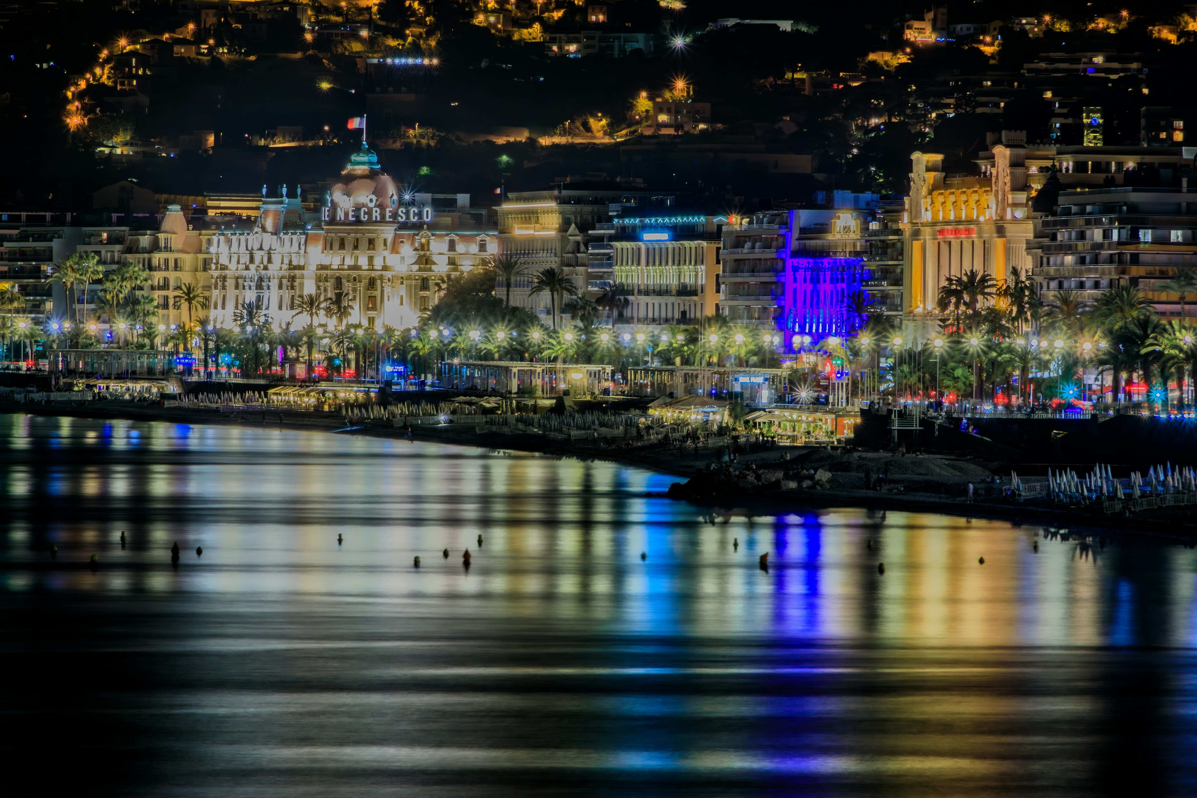 Buildings are illuminated by night along a waterfront road. Their colorful lights are reflected on the web beach in front of them.