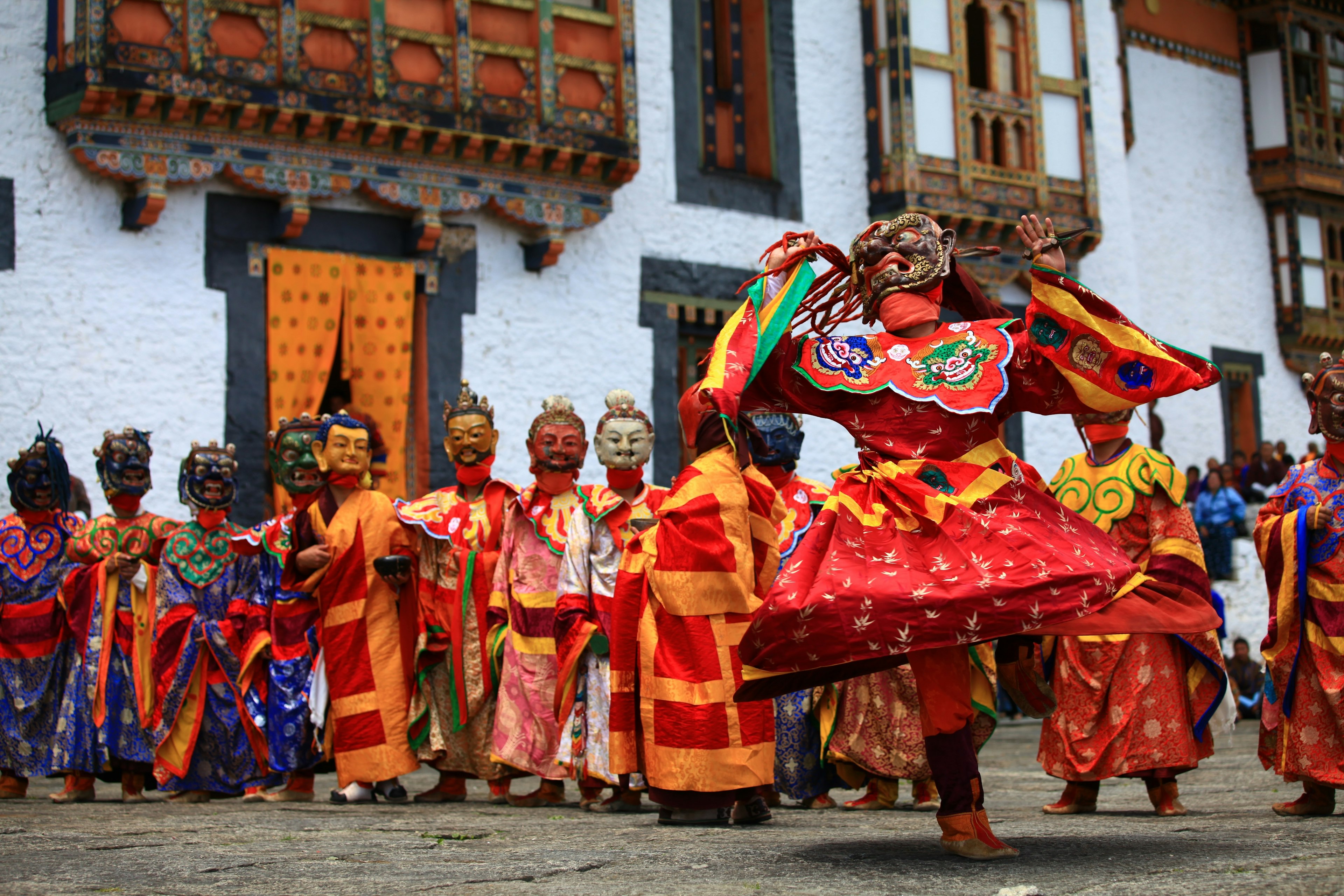 A line of dancers in colorful costumes and masks participates in a festival. A wall with painted wood details and curtains is visible in the back.