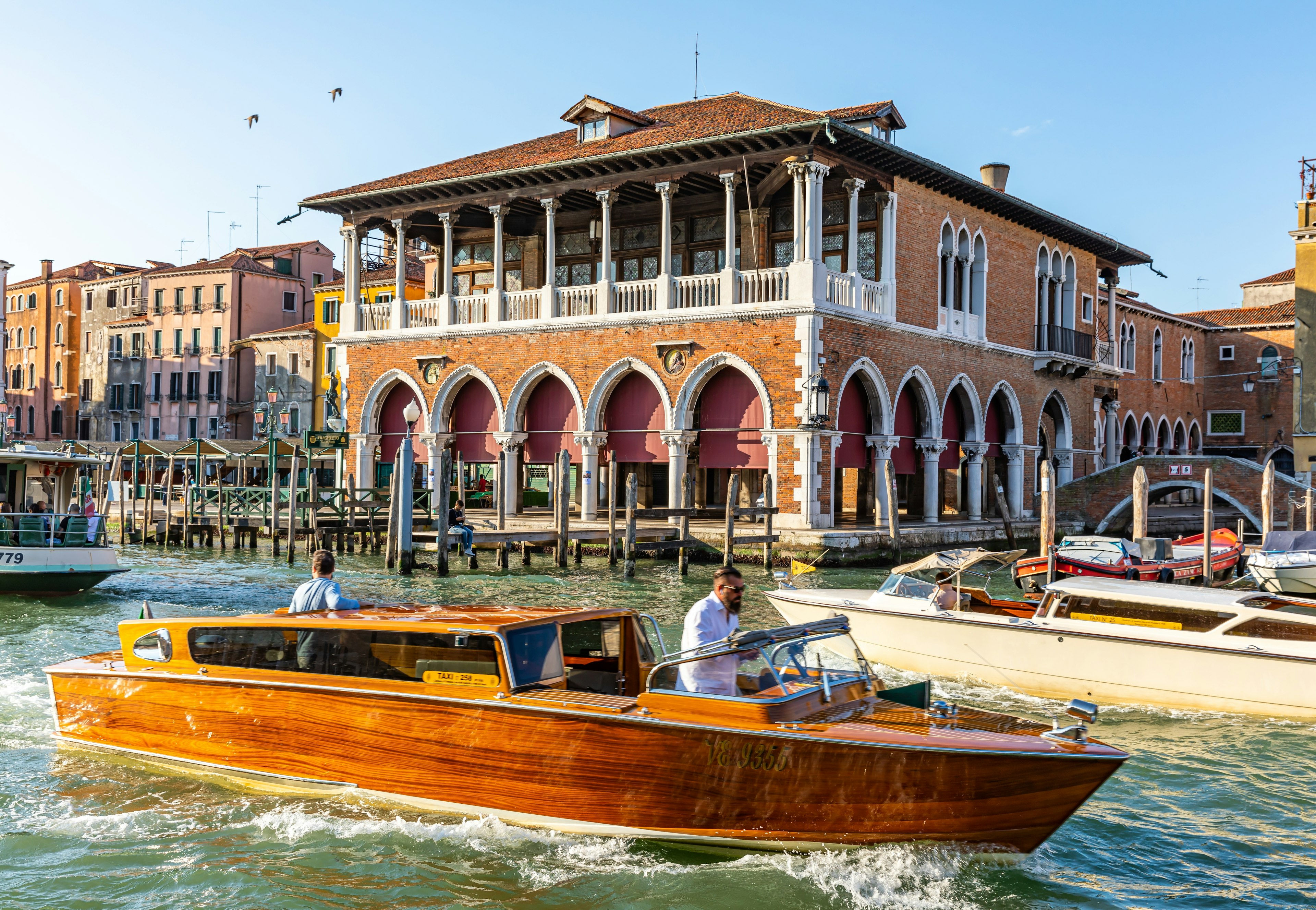 A man in a white shirt pilots a wood-paneled speedboat past an ornate buildings with columns and pointed arches