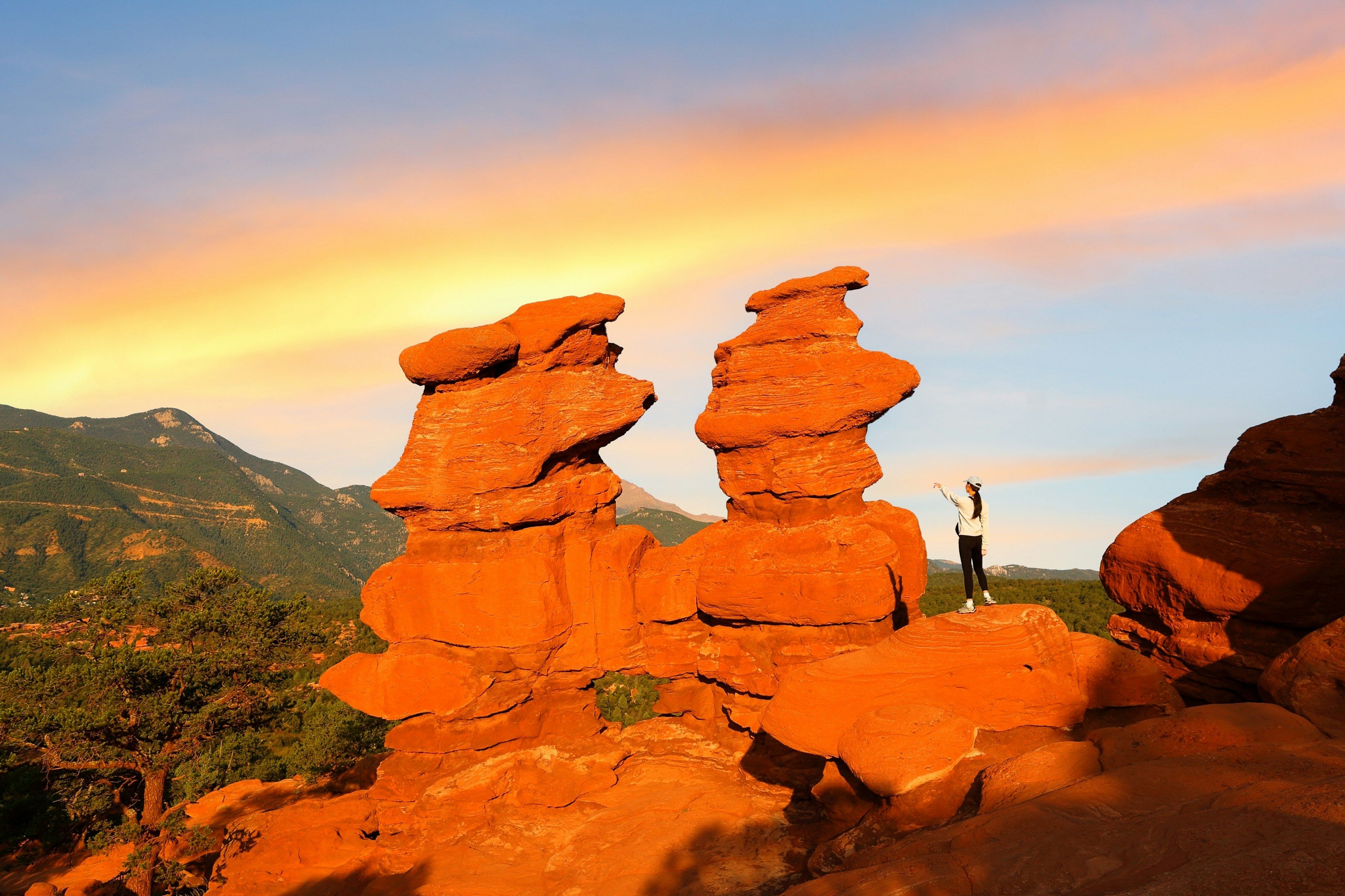 A hiker stands next to two dramatic rock formations at sunset, as the rocks appear red