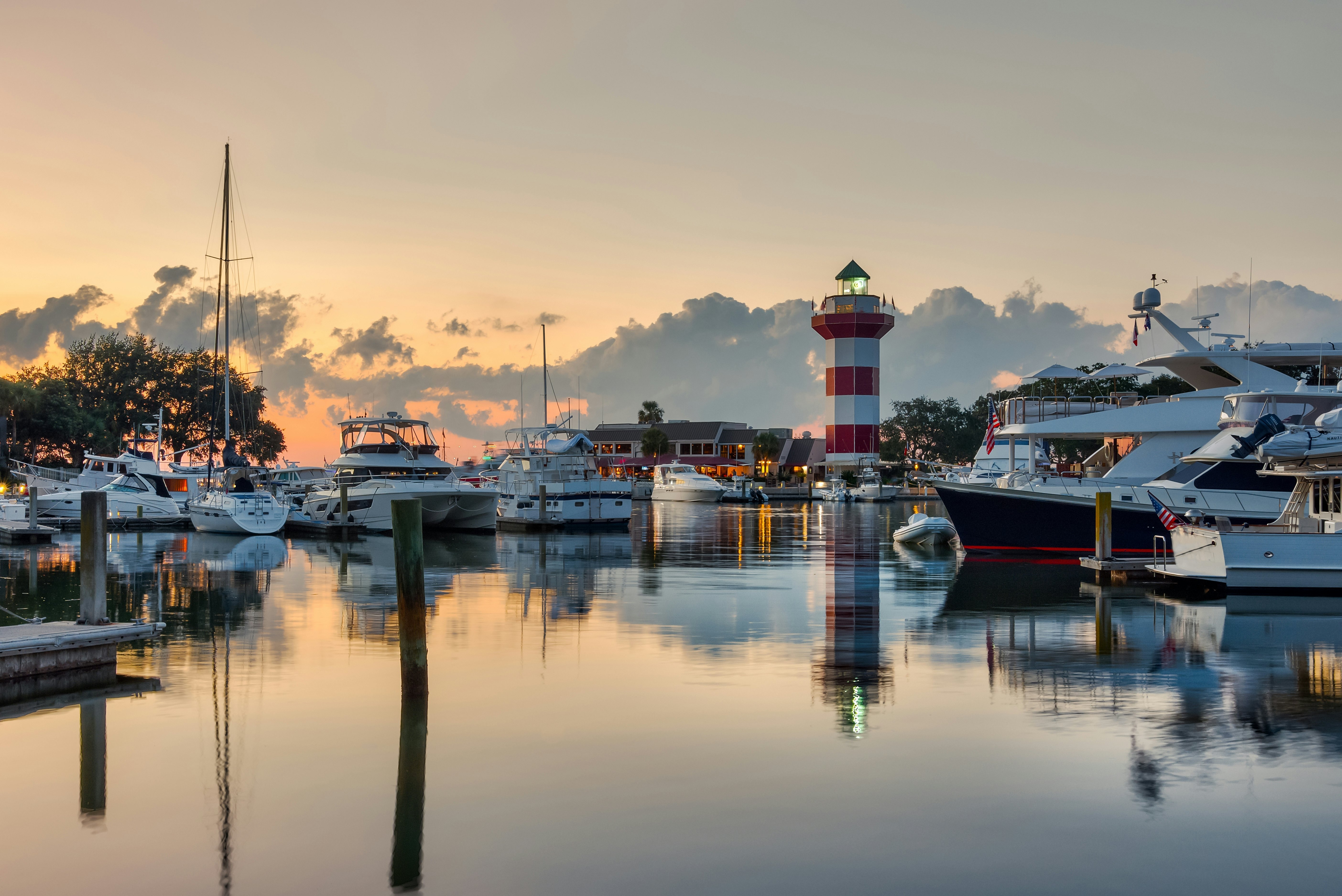 Hilton Head Island and its iconic lighthouse lit up at sunset with a glass like water with reflections of boats and lighthouse