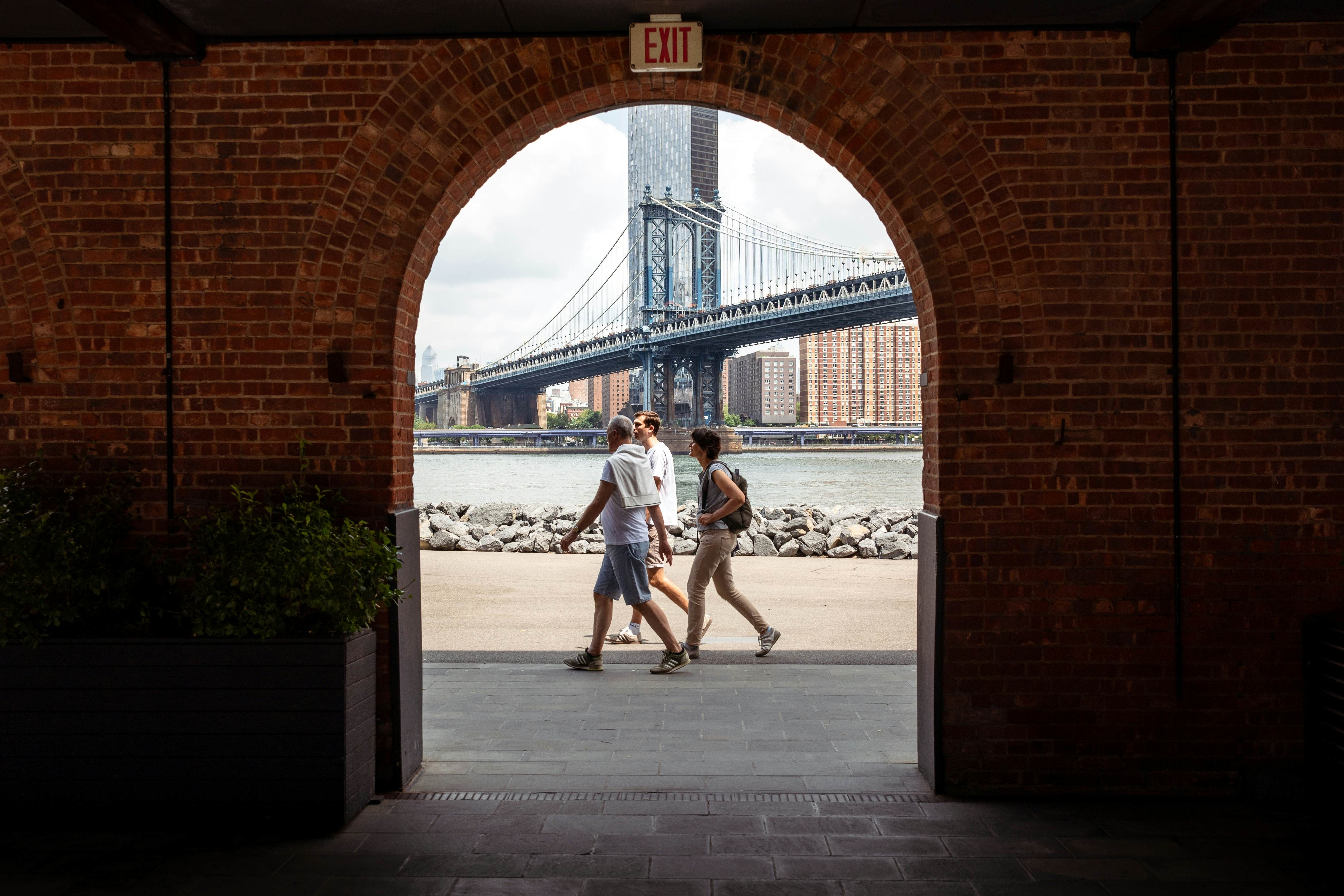 Landscape view of the Manhattan bridge between Brooklyn and Manhattan in New York through a red brick arch with river, rocks and sidewalk in the middle. A family on holiday walks in the middle, License Type: media, Download Time: 2025-01-22T18:19:01.000Z, User: adouglaslott59, Editorial: false, purchase_order: 65050 - Digital Destinations and Articles, job: Future digital articles, client: Future digital articles, other: Ann Douglas Lott