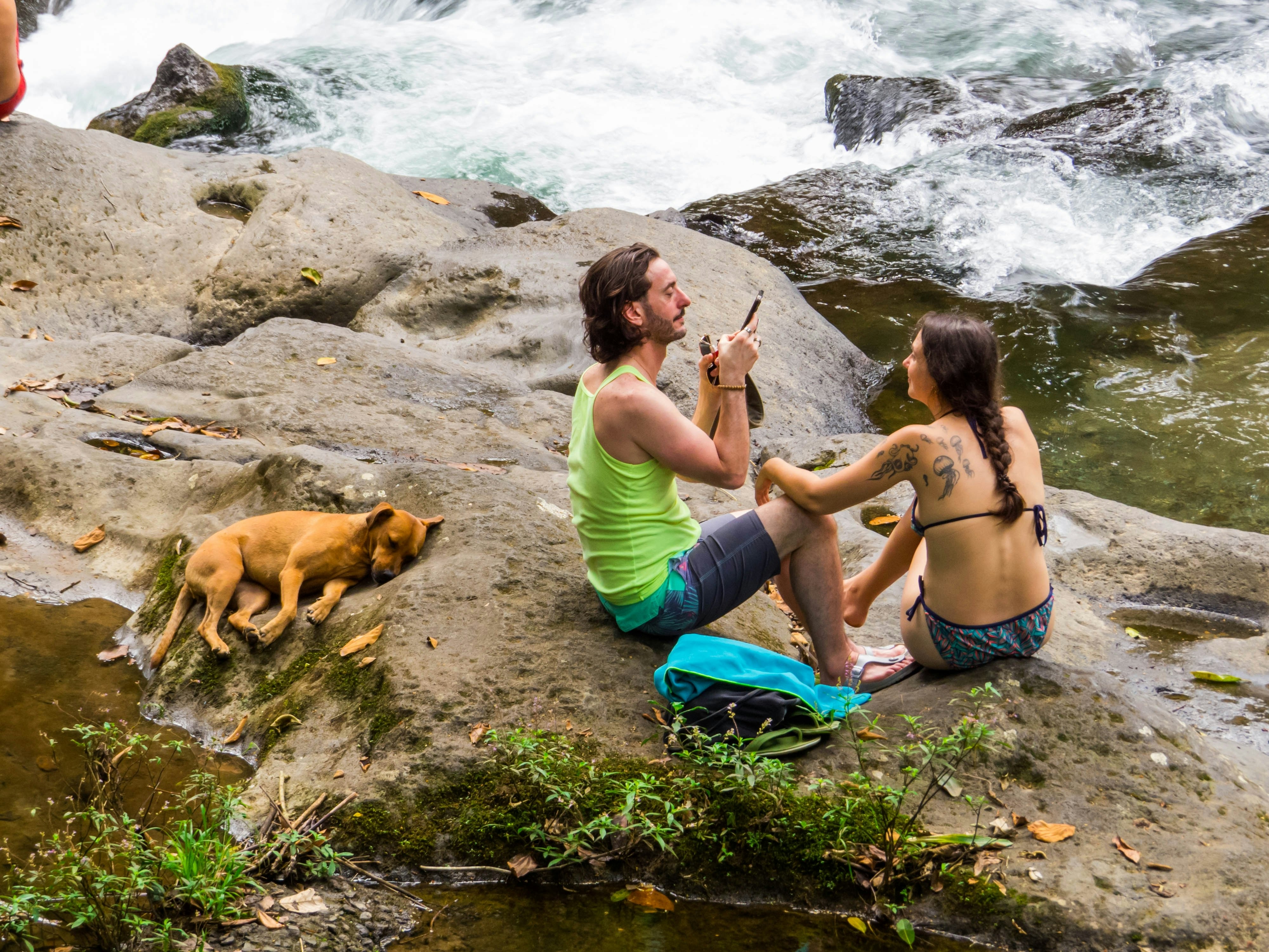 A man holds a phone next to a woman wearing a bikini on rocks by rushing rapids. A dog sleeps on the rocks next to the couple.