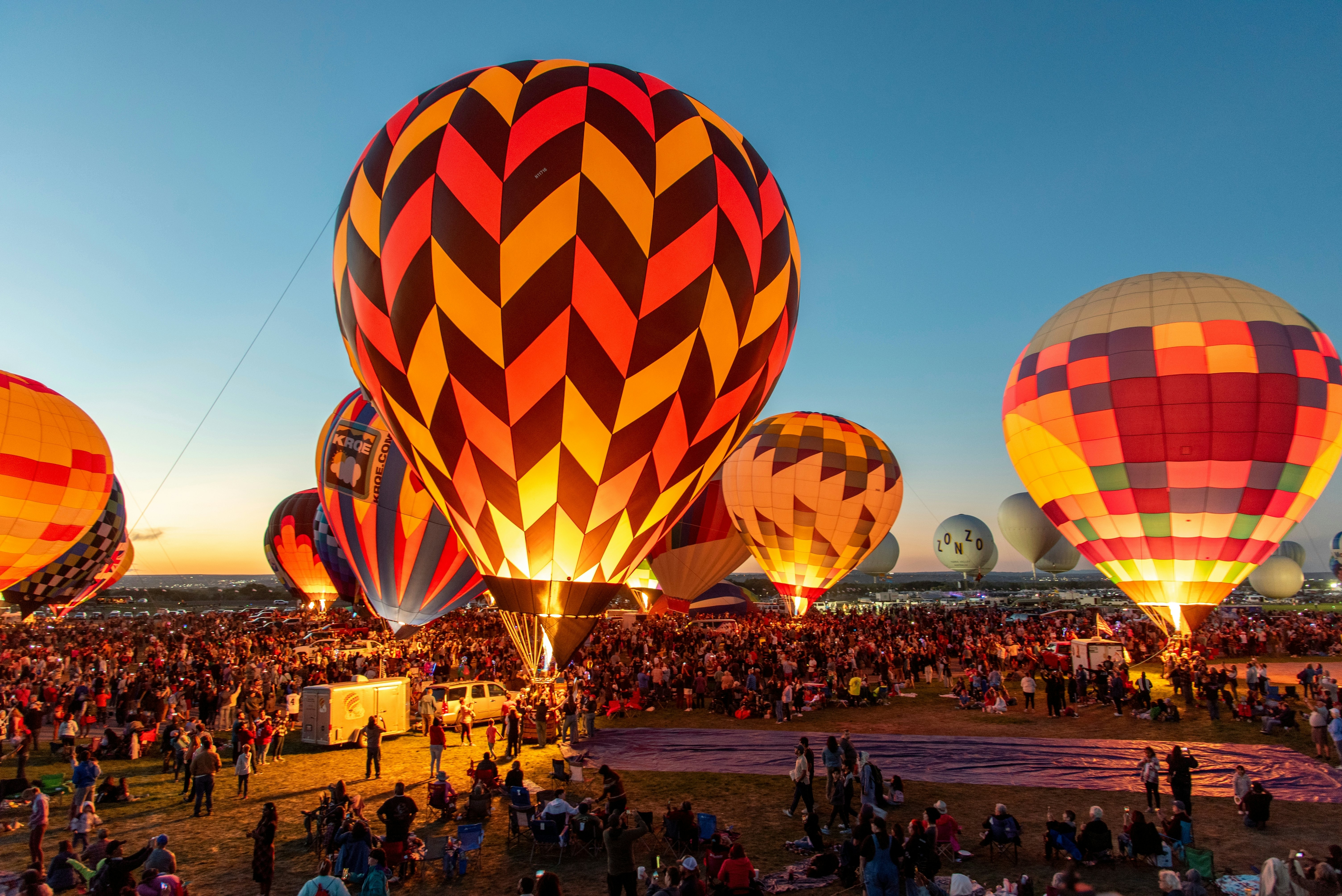 Several hot air balloons in many colors and patterns are inflated by giant flames early one morning. Thousands of people watch from the ground.