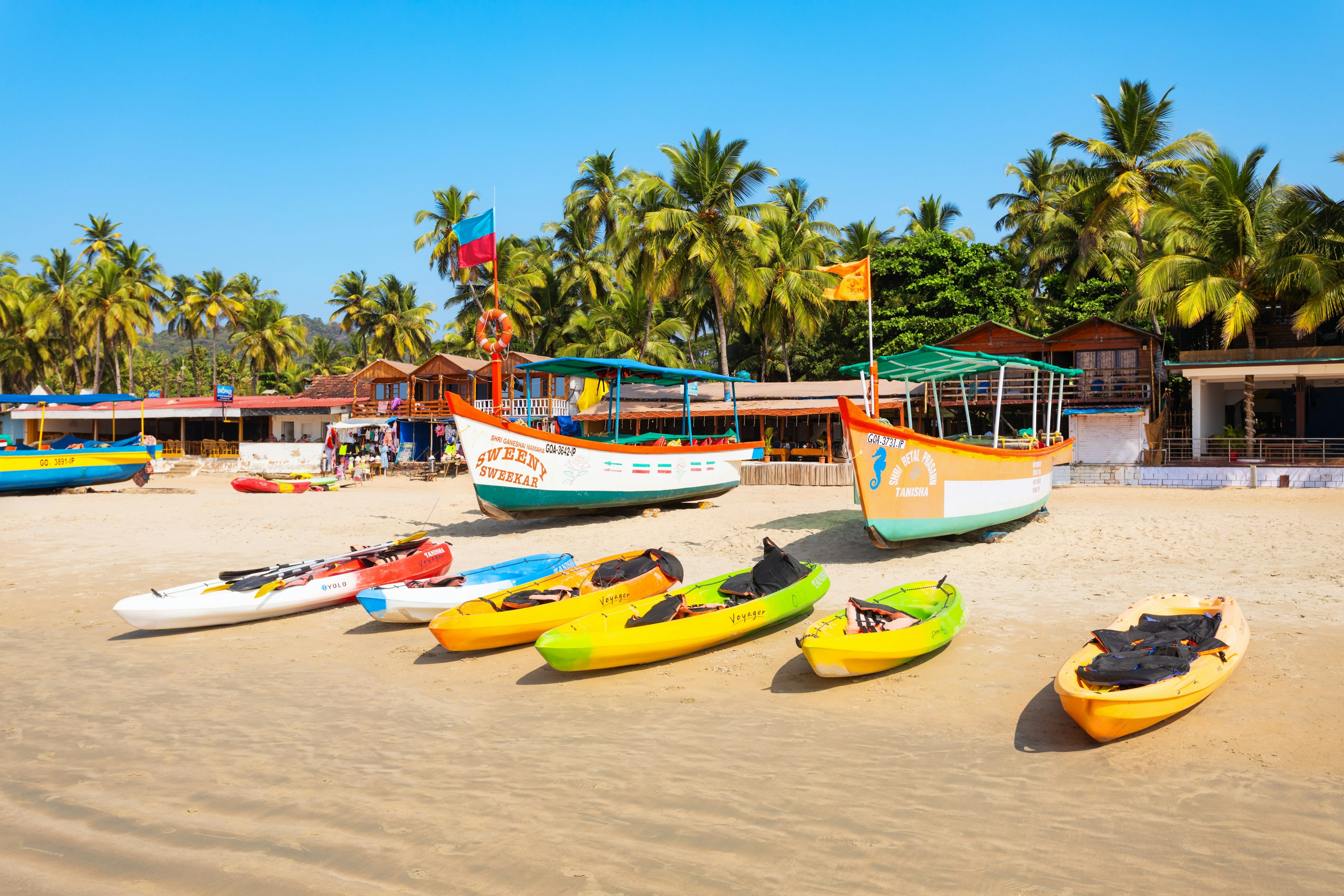 Colorful kayaks, fishing boats and other water craft are lined up on a beach. A row of buildings is shaded by palm trees behind the boats.