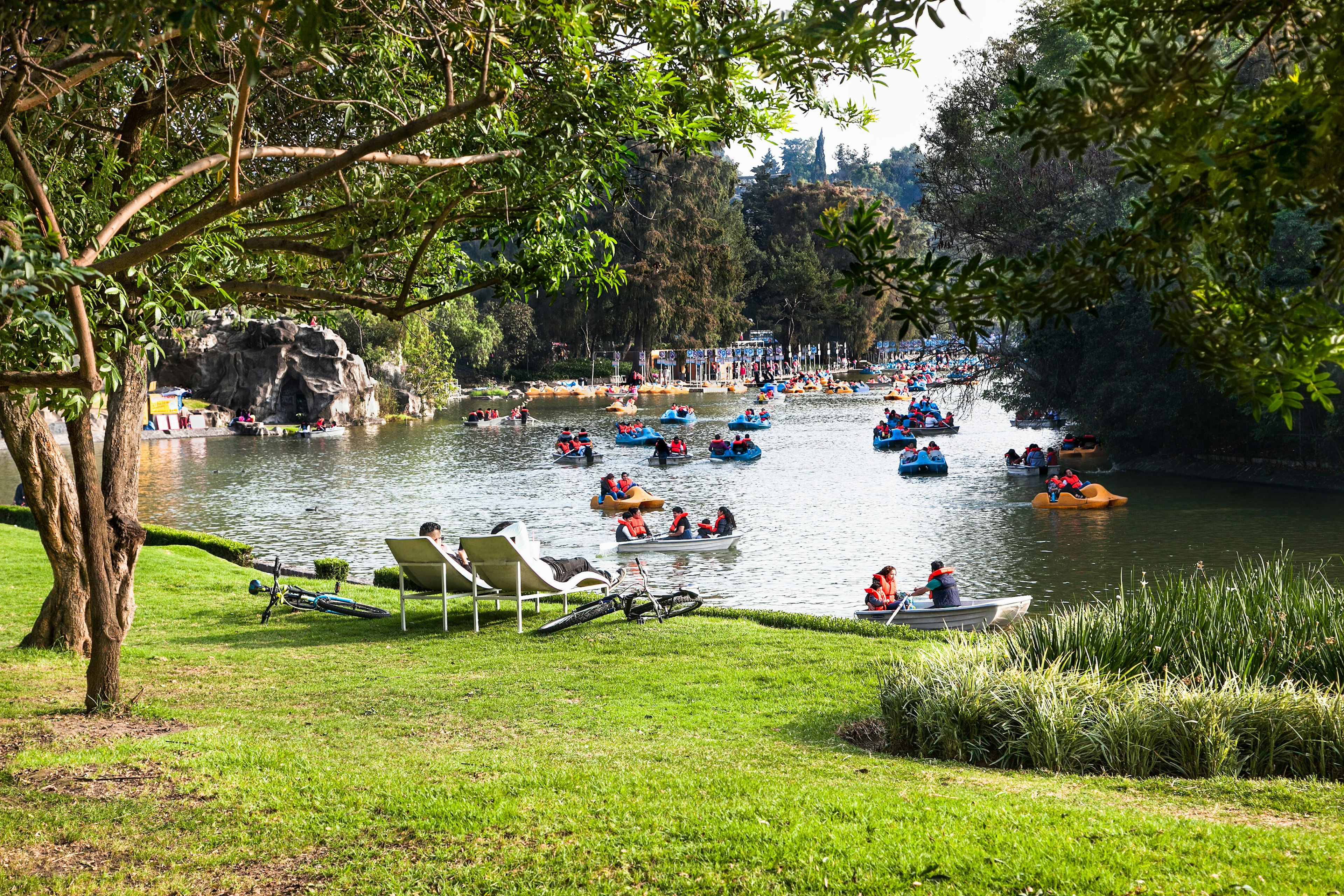 A lake in a city park is pictured. People are in rowboats and paddle boats on the water, while others sit in loungers on a lawn looking out at the water.