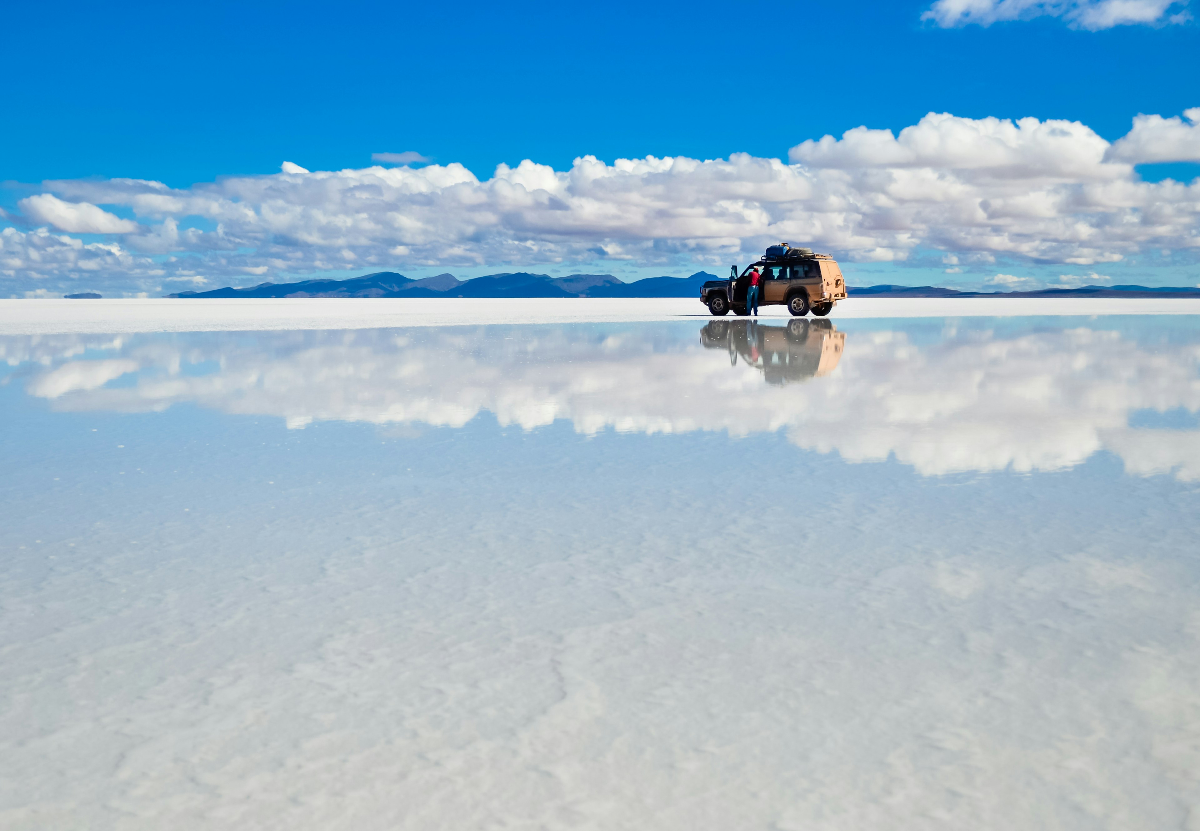 Vast white salt flats stretch into the distance, meeting the blue sky and far hills, with a 4WD reflected in the surface water