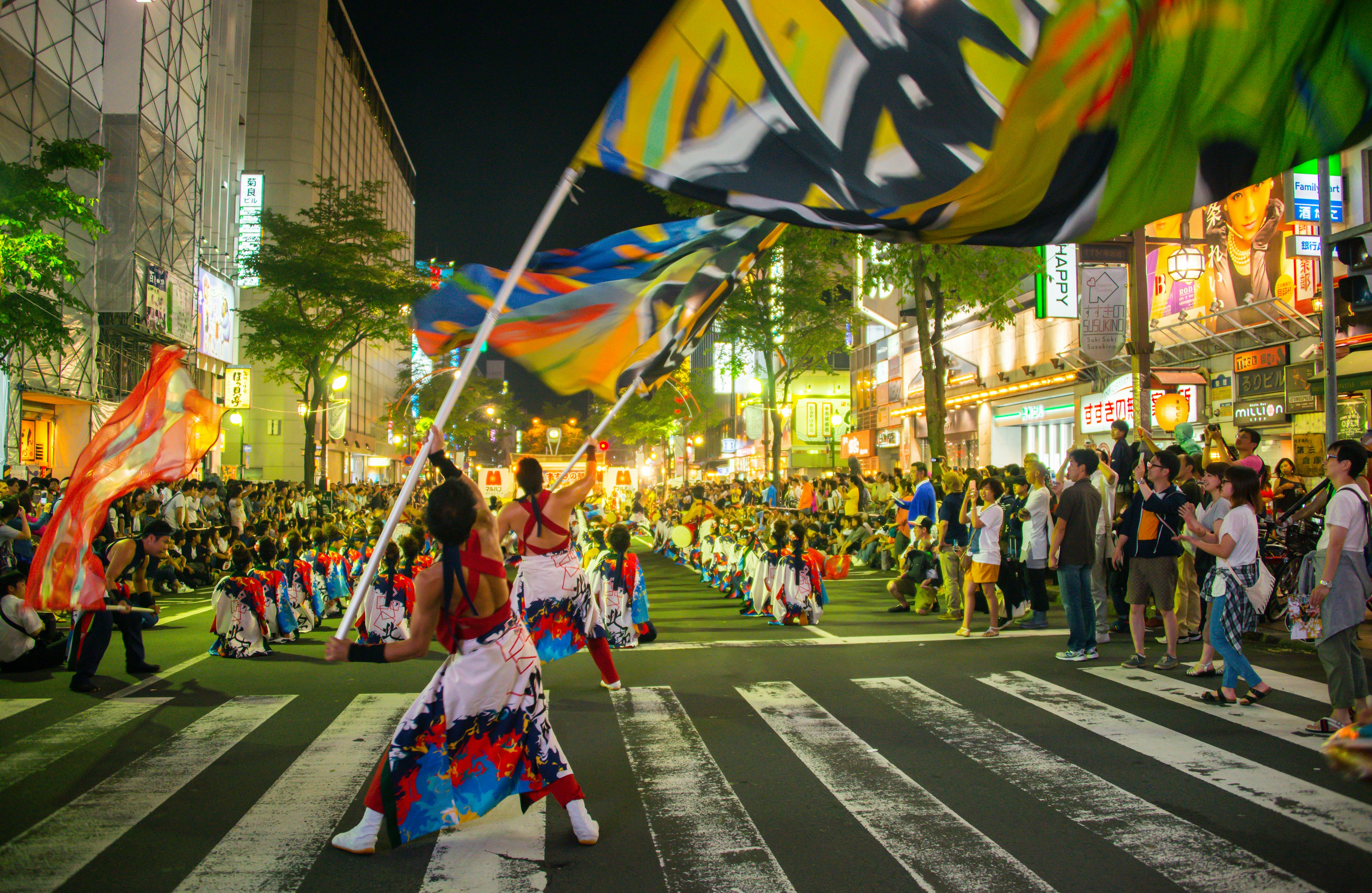 People in costume wave festive flags during a festival on a city street at night. Crowds of onlookers admire the performers.