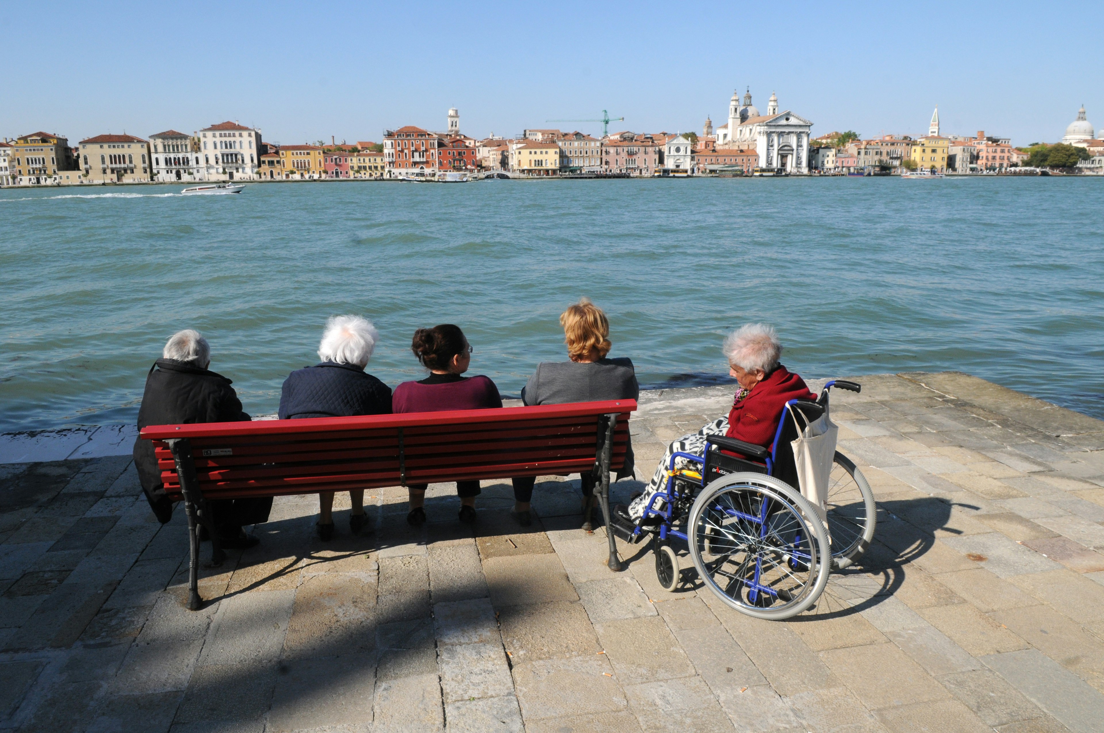 Four people sit on a bench next to an elderly woman in a wheelchair on the waterfront, looking at out a canal and buildings on the shore in the distance