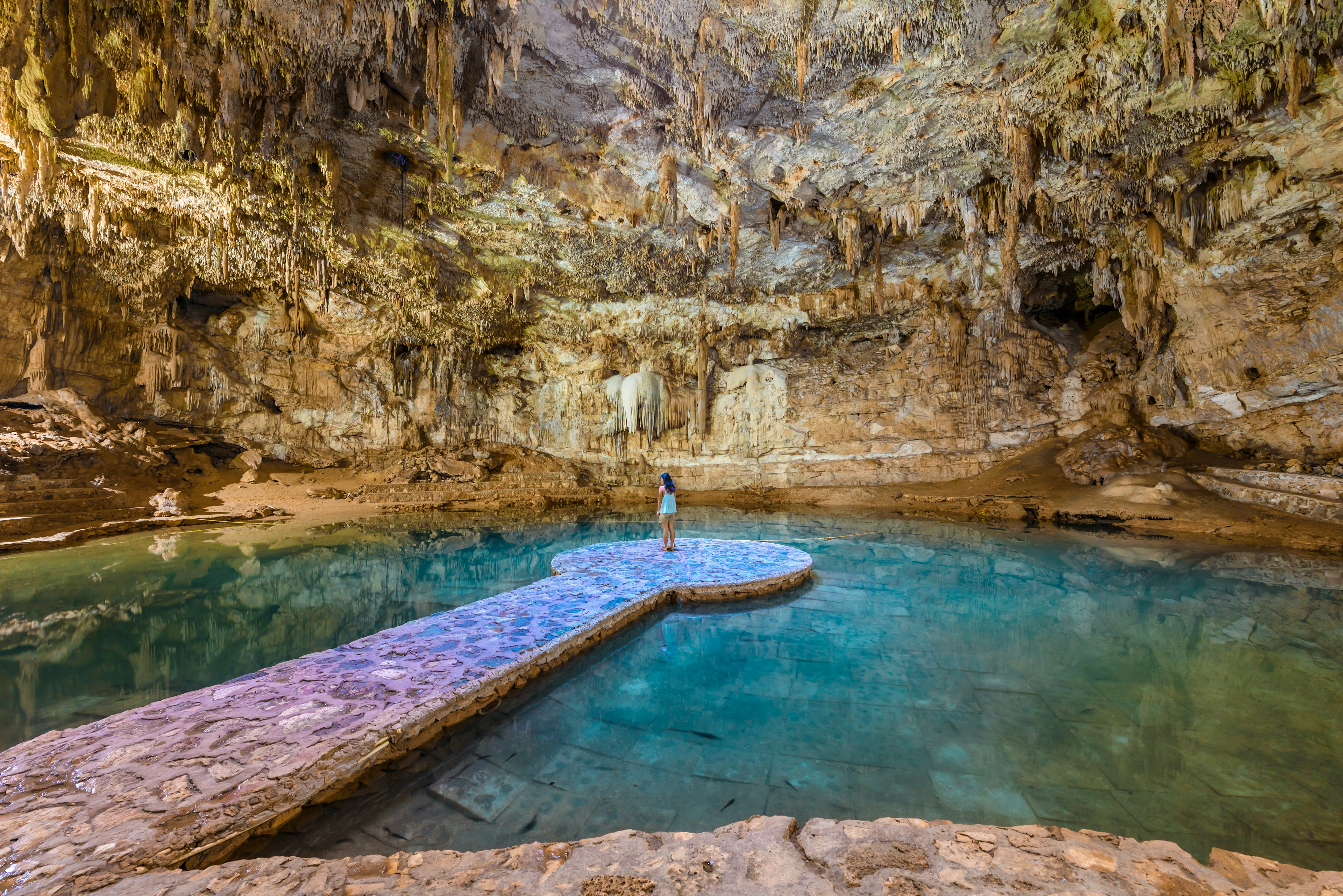 A woman stands at the end of a platform in a pool in a cave, with stalactites covering the cave dome. A beam of lights illuminates the underground scene.