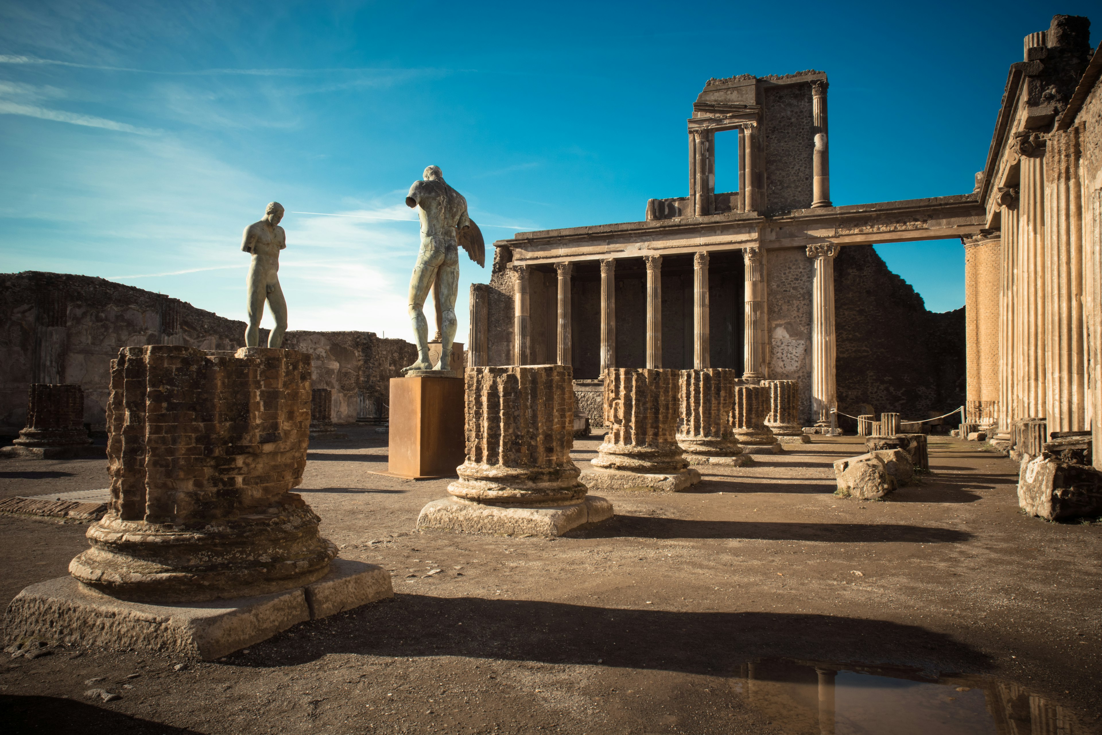 Ruined columns and monumental statues in a courtyard surrounded by other columns, under a blue sky with dramatic shadows