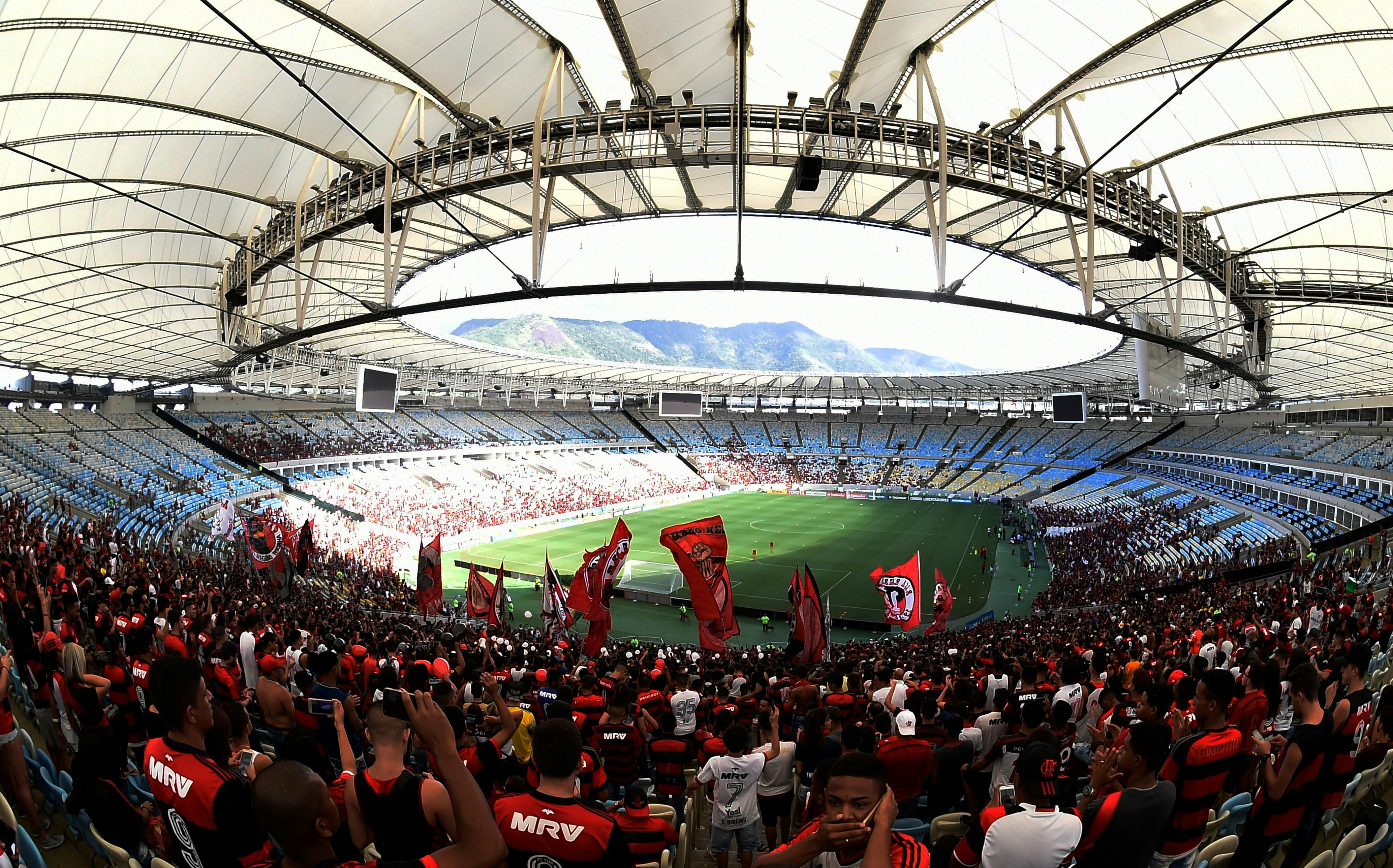 A wide view of fans in red home-team jerseys and holding flags cheering on a match at a giant stadium. An oculus through the partially covered roof lets sunlight onto the field.