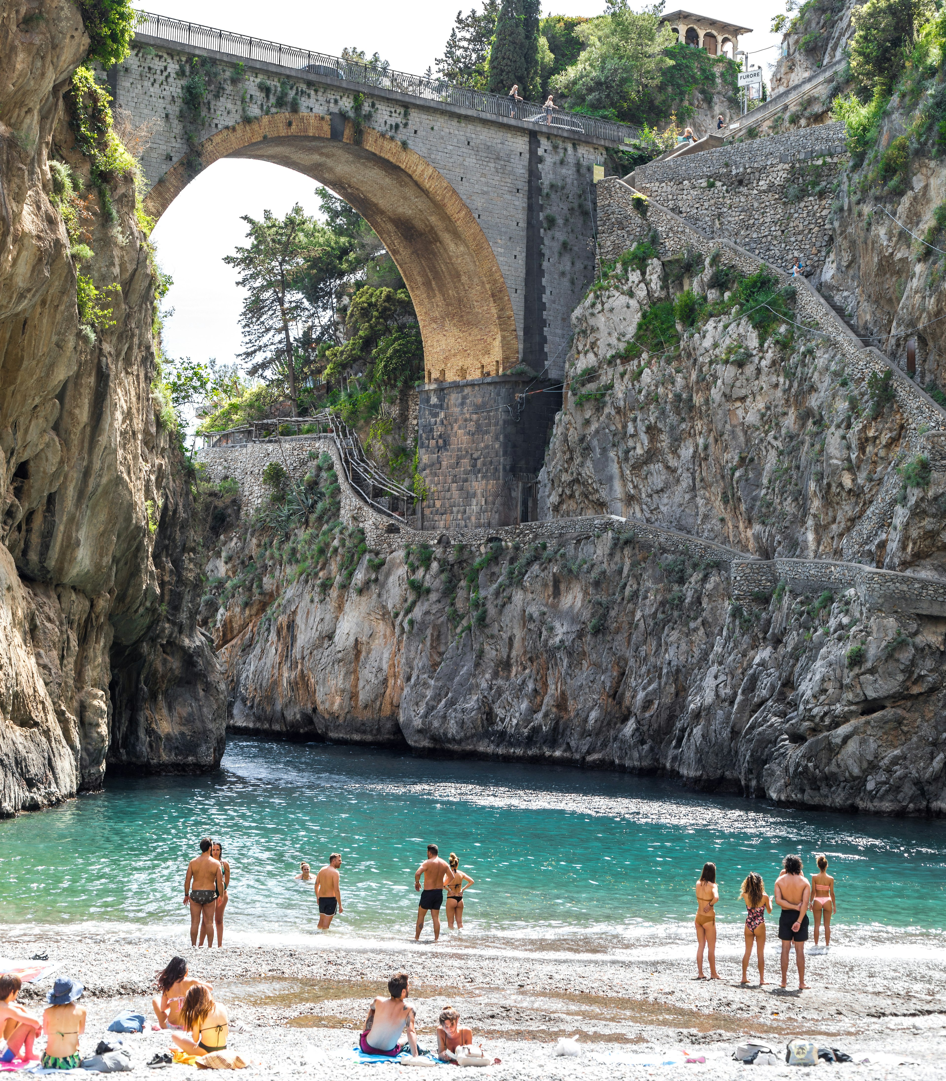 People swimming and relaxing at the beach with clear blue water and a bridge overhead.