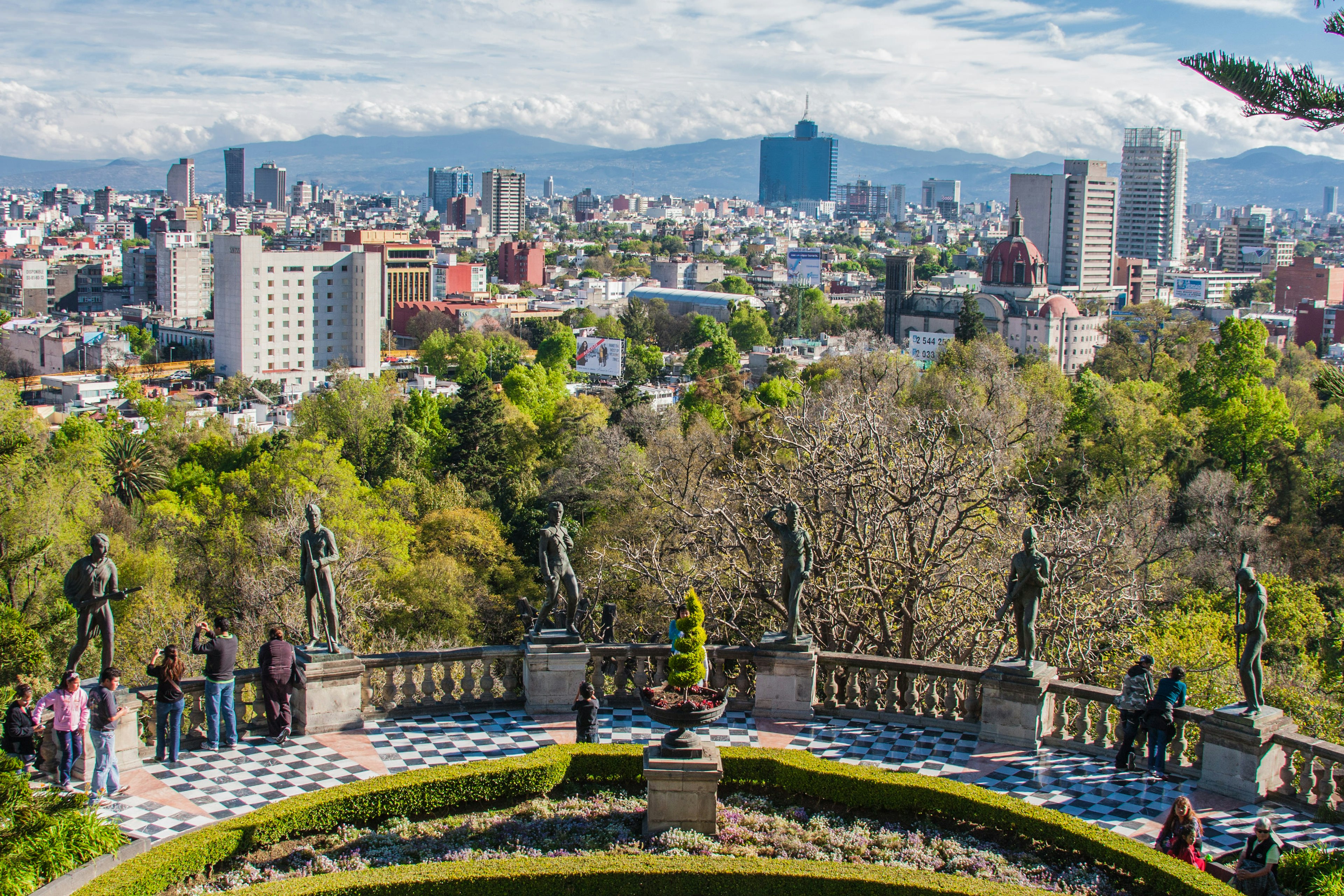 A viewpoint in parkland with people gazing out between sculptures at the view of the city beyond