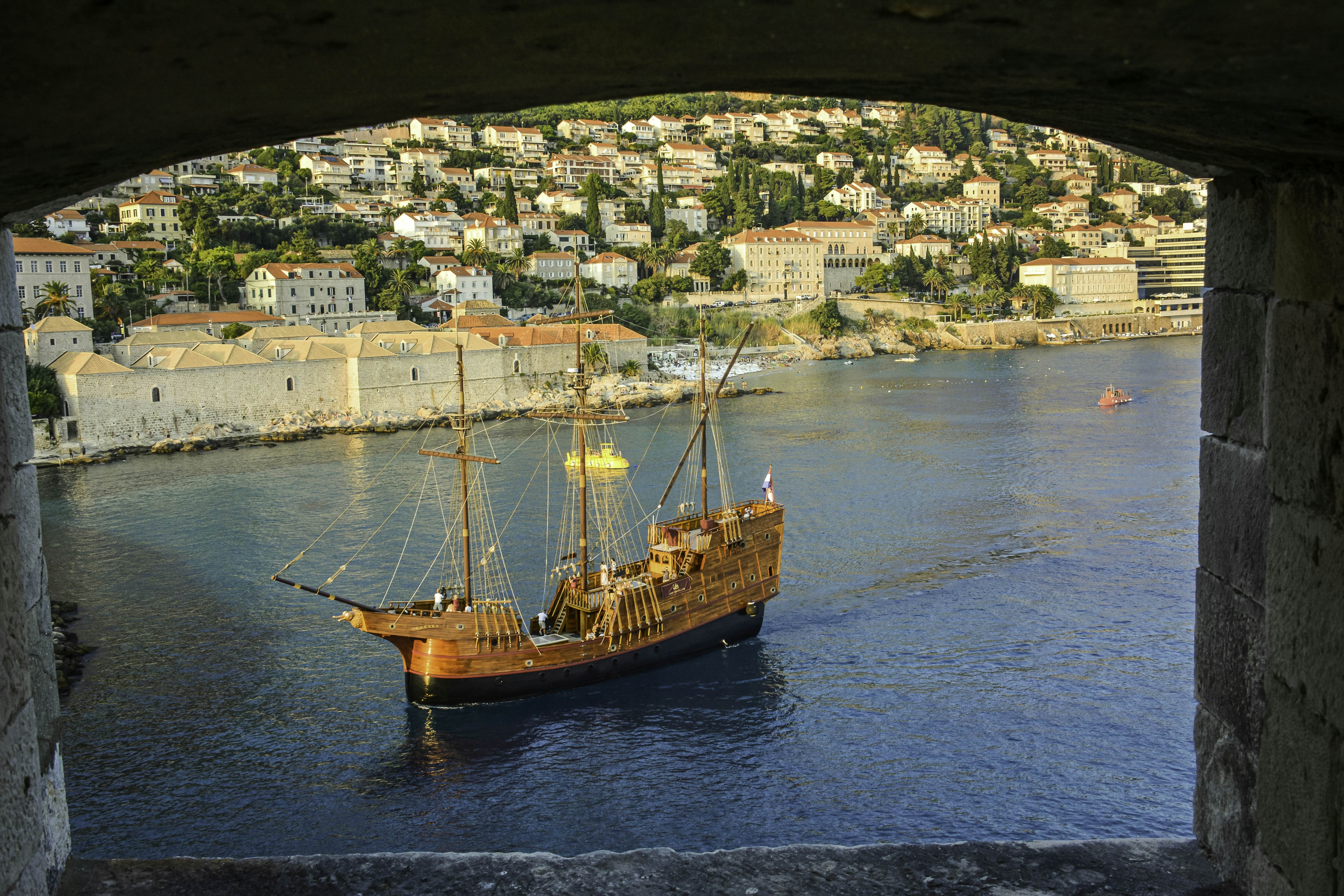 A three-masted sailing vessel in a city harbor glowing in the sun