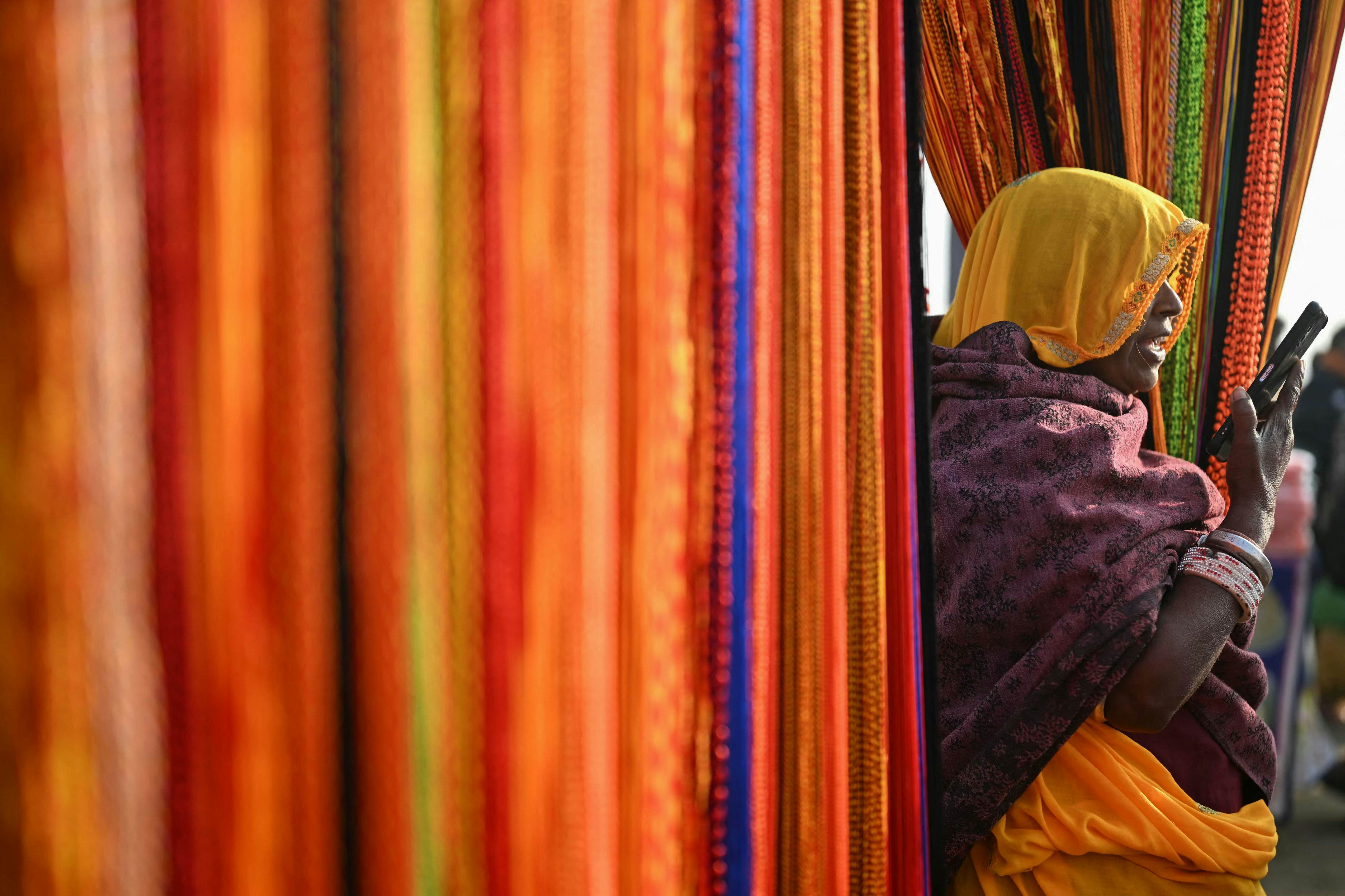 A woman with a head covering speaks into a phone at a stall draped with colorful fabrics.